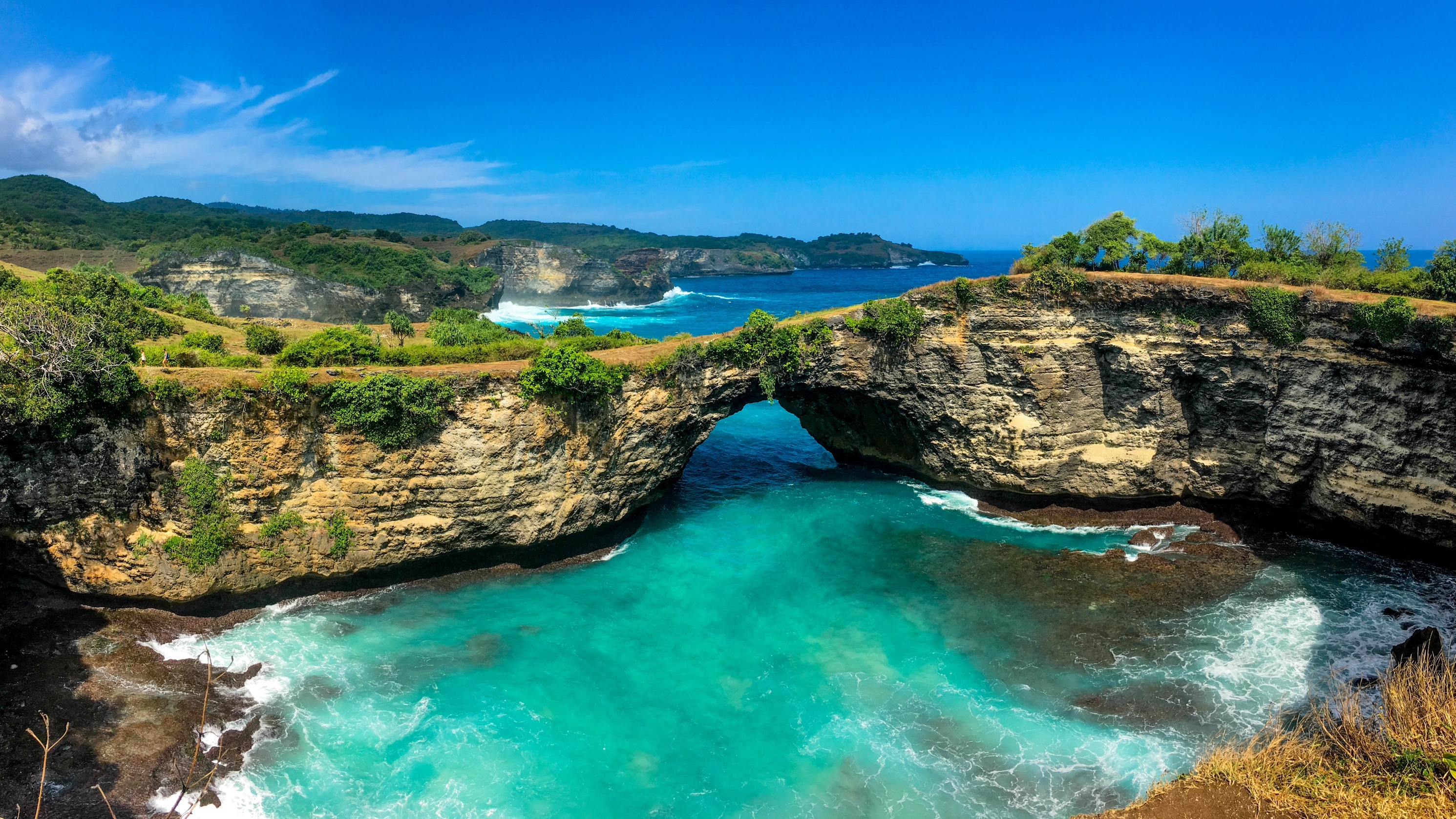 A stunning, high-angle panoramic view of Broken Beach (Pasih Uug) on Nusa Penida, Indonesia, under a brilliant, clear blue sky. The focal point is a massive natural limestone arch that forms a bridge over the water, connecting the rugged cliffs. This arch encircles a spectacular, circular cove filled with vibrant, glowing turquoise water. White waves crash at the base of the cliffs where the ocean flows into the lagoon. The surrounding cliffs are topped with green and brown vegetation, and the deep blue of the open ocean is visible in the background.
