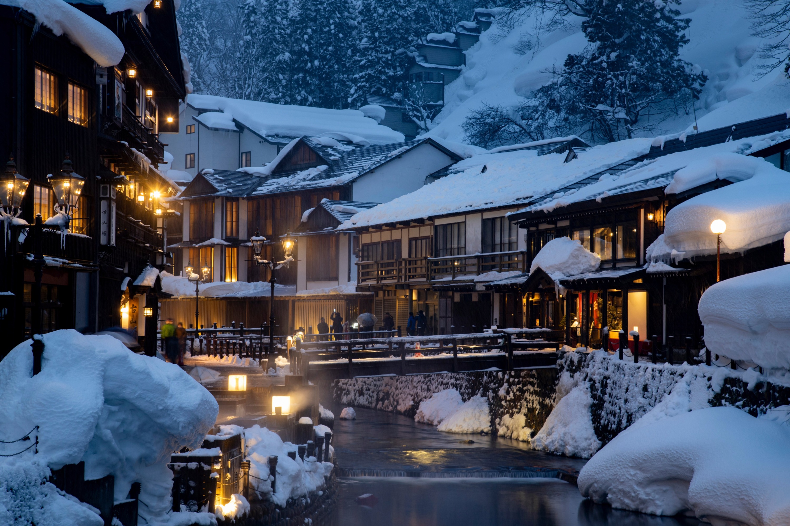 A magical, snowy evening scene at Ginzan Onsen in Yamagata, Japan. A narrow, pedestrian-only street is lined on both sides by historic, multi-story wooden ryokans (inns), their architecture reminiscent of the Taisho period. A small river flows through the center of the walkway, crossed by several small, snow-covered footbridges.