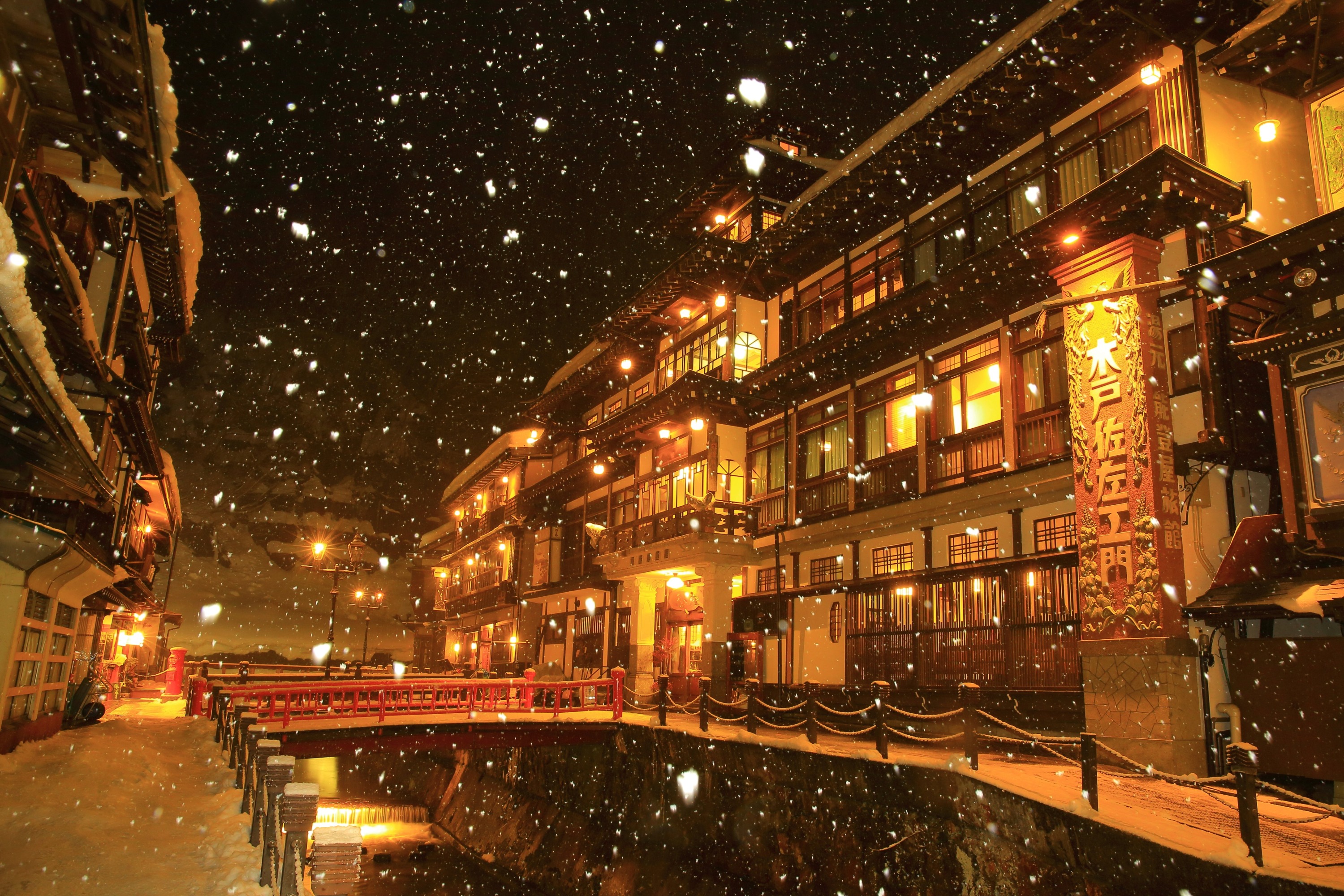 A breathtaking, enchanting winter night scene of Ginzan Onsen in Obanazawa, Yamagata, Japan. The central Ginzan River flows calmly through the snow-covered village, reflecting the warm, golden light emanating from the traditional wooden ryokans (inns) that line both banks. These multi-story inns, with their distinctive architecture and intricate wooden details, are completely blanketed in a thick layer of fresh, white snow on their roofs and balconies. Ornate gas street lamps glow brightly, casting a soft yellow light that highlights the falling snowflakes and creates a magical, ethereal atmosphere. Several snow-covered footbridges arch gracefully over the river, connecting the two sides of the charming, historic street. The overall impression is one of serene beauty, warmth, and nostalgic charm amidst a pristine winter wonderland.