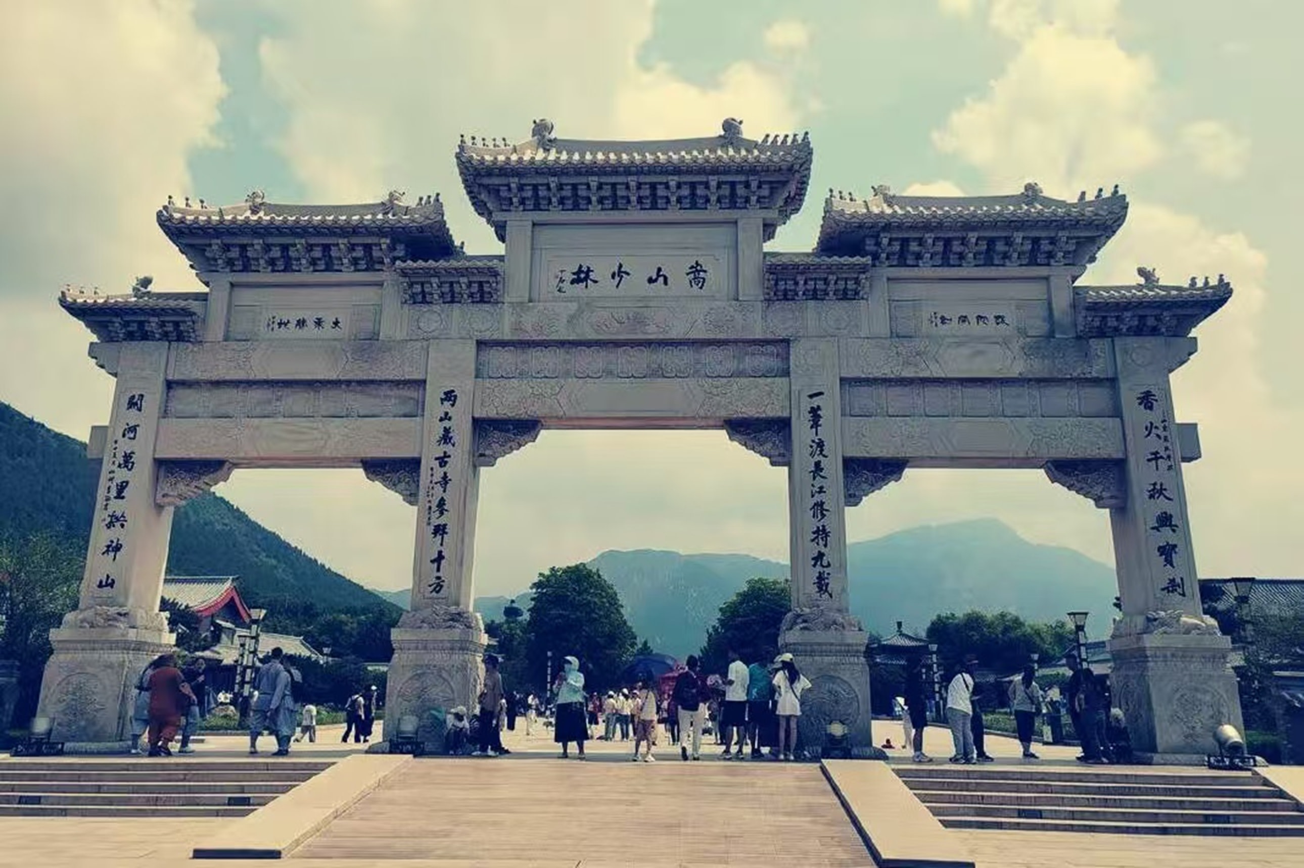 這是一座宏偉的四柱三門石牌坊，正中題有 “嵩山少林” 四字，兩側立柱刻有對仗楹聯，雕飾精美繁複。牌坊後是連綿的青山與蔥鬱樹木，遊人往來其間，既顯古建的雄渾氣勢，也透著景區的菸火氣