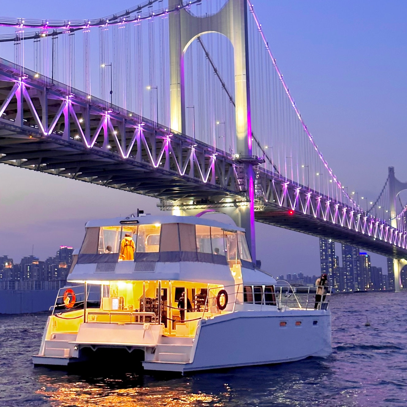 A yacht beneath an illuminated suspension bridge in Busan at dusk.