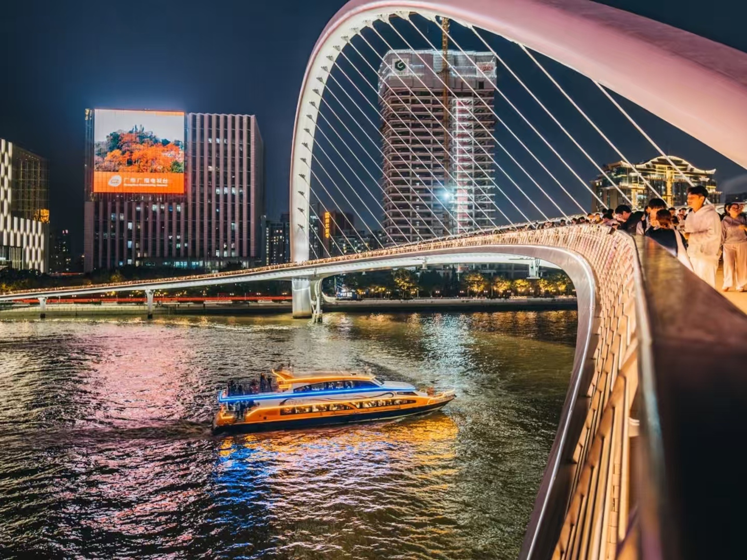 海心橋下,遊船泛著藍黃燈光駛過珠江,橋面行人駐足觀景,背景建築大屏映著秋景圖,兩岸燈光與江水倒影交織,夜遊的熱鬧與城市夜景相融 海心橋下,遊船泛著藍黃燈光駛過珠江,橋面行人駐足觀景,背景建築大屏映著秋景圖,兩岸燈光與江水倒影交織,夜遊的熱鬧與城市夜景相融