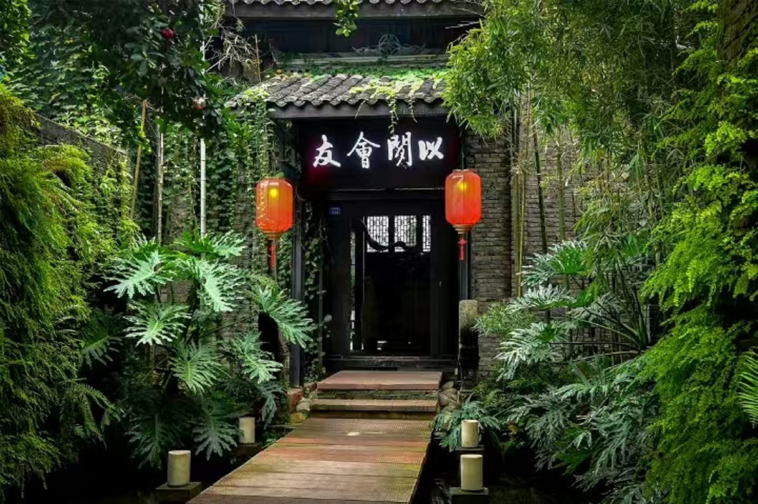 古意盎然的院落入口，青磚灰瓦間爬滿翠綠藤蔓，兩盞紅燈籠映照出 “以閣會友” 的牌匾，木質棧道穿梭於蔥鬱綠植與潺潺流水之間，彷彿一處藏於都市的雅緻秘境，正靜待知音造訪