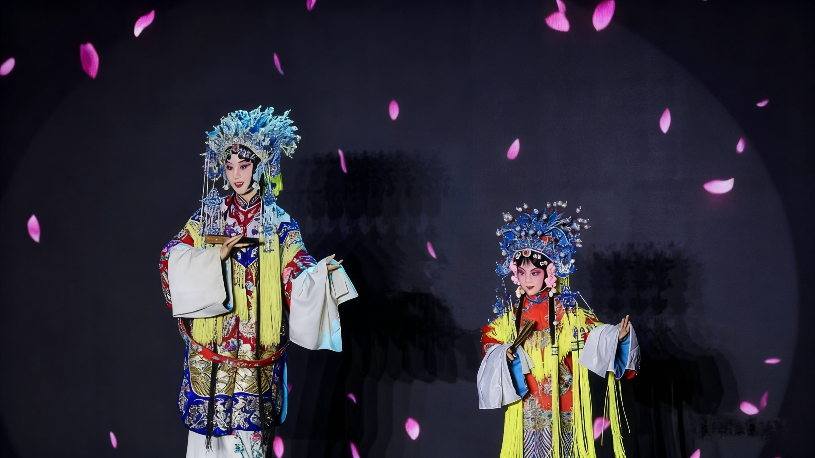 兩位京劇旦角演員身著華麗的戲服,搭配藍色的鳳冠,格外亮眼。背景的花瓣與深色幕布營造出浪漫的氛圍,似乎在演繹一段宮廷中的唯美故事,每一個動作都透著旦角的溫婉與典雅 兩位京劇旦角演員身著華麗的戲服,搭配藍色的鳳冠,格外亮眼。背景的花瓣與深色幕布營造出浪漫的氛圍,似乎在演繹一段宮廷中的唯美故事,每一個動作都透著旦角的溫婉與典雅