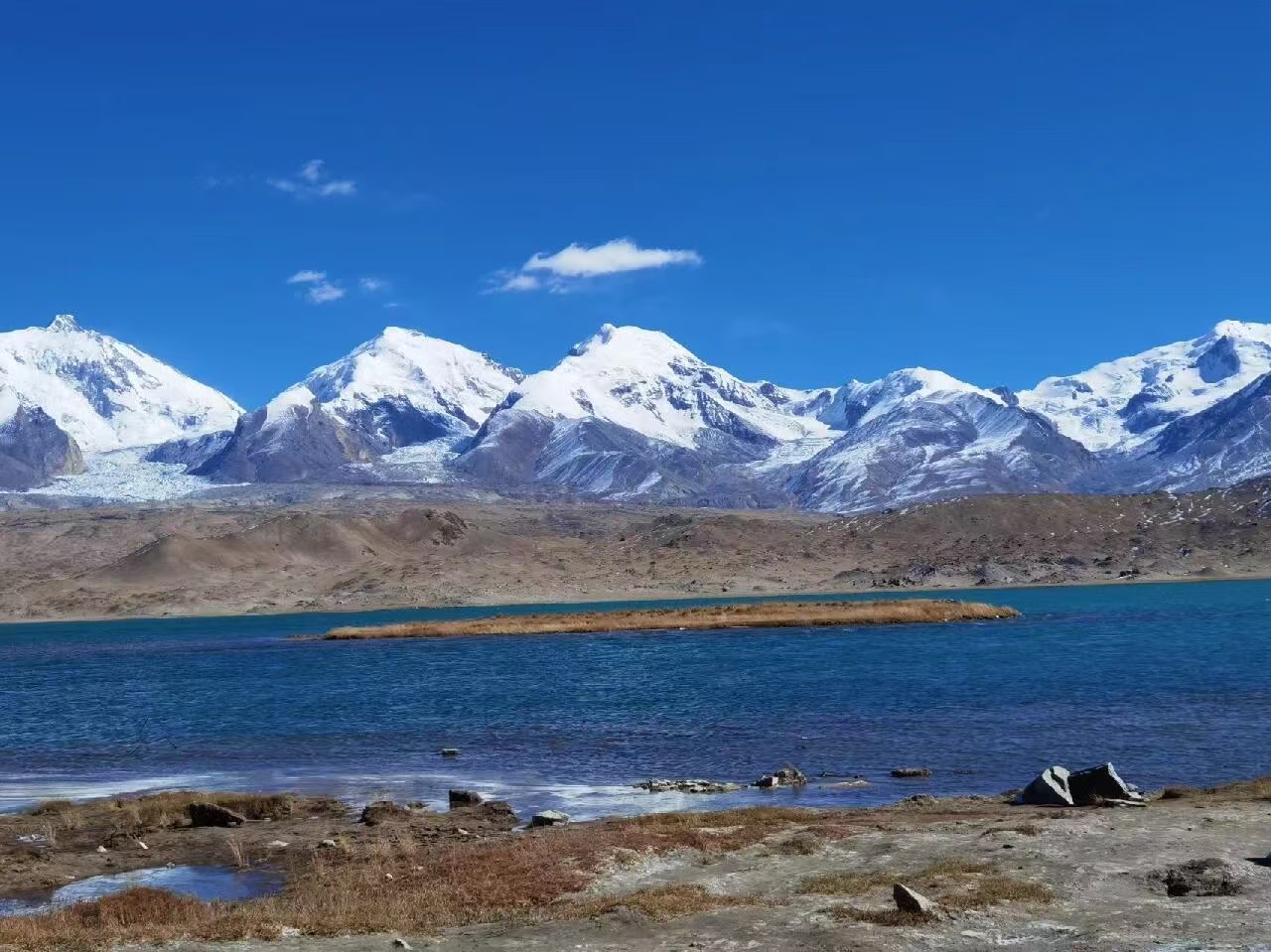 帕米爾高原的湖泊旁，連綿的雪山群峰在湛藍的天空下一字排開，雪峰潔白耀眼。湖水呈現出深邃的藍調，岸邊的荒原與岩石點綴其間，盡顯高原的雄渾與遼闊