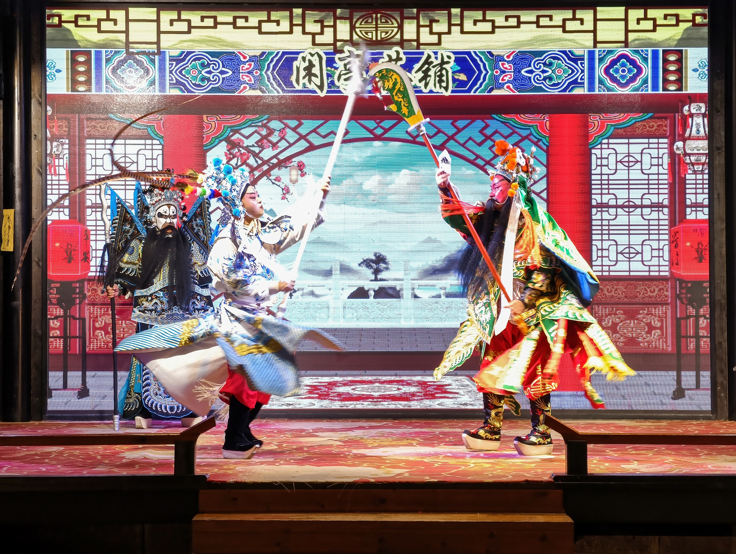 身著精緻戲服，手持兵器，在 “閑亭茶鋪” 的佈景前激烈對打，舞臺背景的山水畫面與傳統建築元素，為這場武戲增添了濃郁的古典氛圍