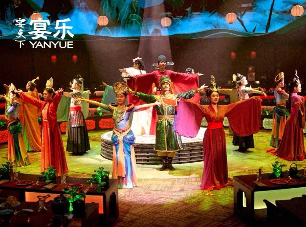 舞臺上演員身著唐風華服，色彩明豔的襦裙、官袍搭配精緻頭飾，舒展衣袖擺出大氣舒展的造型；背景是 360 度環繞的國風大屏，水墨山水與宮燈元素交織，襯得整個場景既有盛唐宴樂的雍容，又帶現代舞美的絢爛。