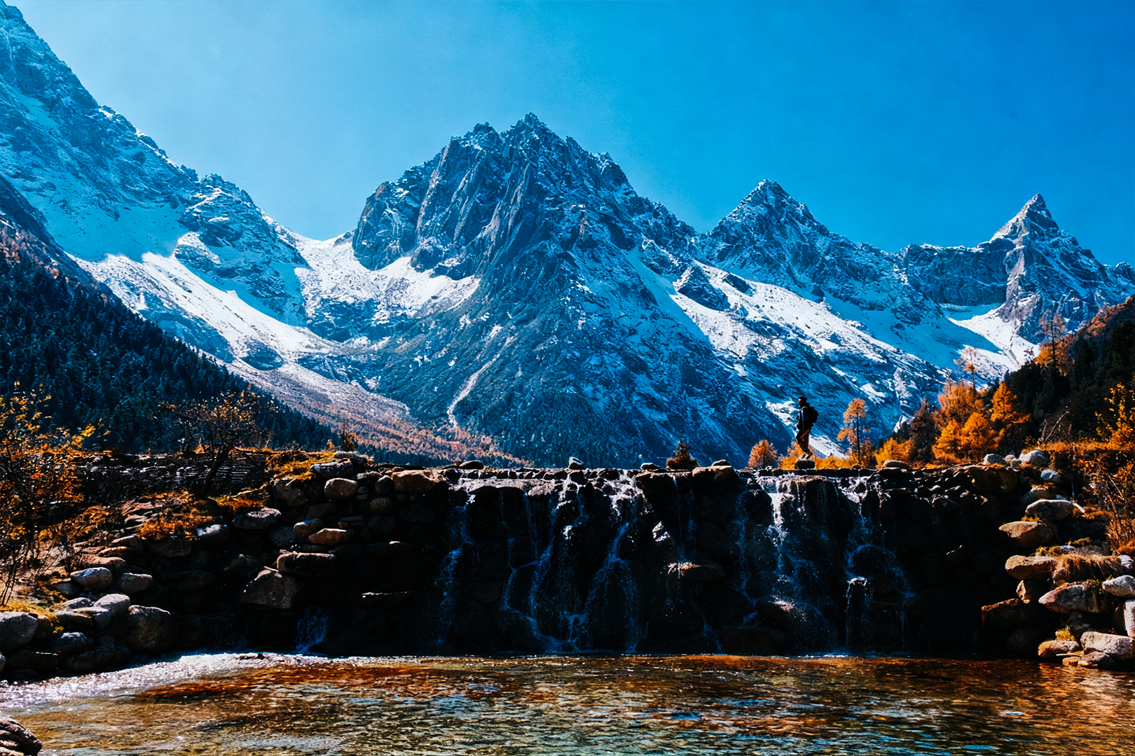 瀑布如銀簾垂落，在岩石間濺起晶瑩的水花；遠處雪山巍峨，在藍天的映襯下更顯冷峻