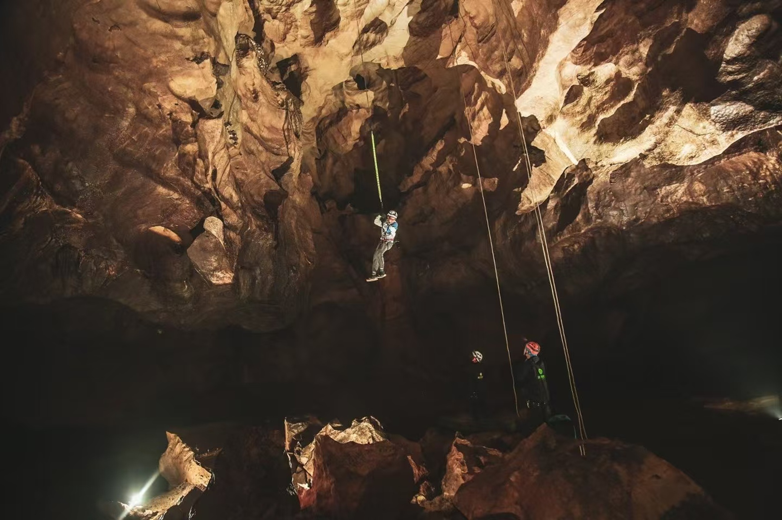 探險者在溶洞內繩降而下,巖壁紋理粗獷,暖黃燈光勾勒出洞穴縱深,下方隊友待命,盡顯地心探險的刺激與專業 探險者在溶洞內繩降而下,巖壁紋理粗獷,暖黃燈光勾勒出洞穴縱深,下方隊友待命,盡顯地心探險的刺激與專業