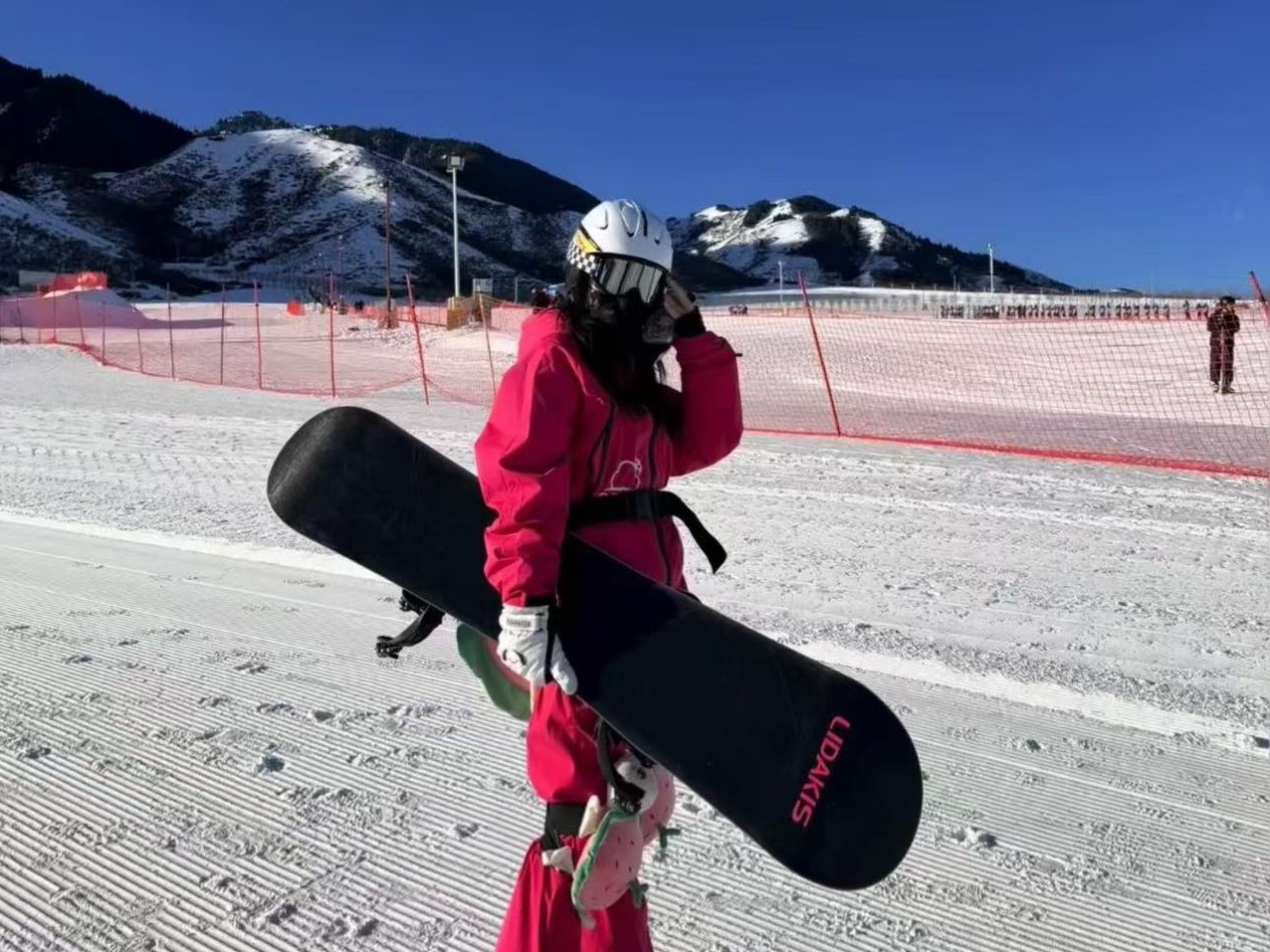 晴日雪場裡，身著亮紅色連體滑雪服的雪友站立雪道，手持 LIDAKIS 單板，佩戴白頭盔與護目鏡，搭配可愛護臀護具。背景是覆雪山巒與防護網，藍天澄澈，整體氛圍清爽亮眼，盡顯單板滑雪潮流穿搭感