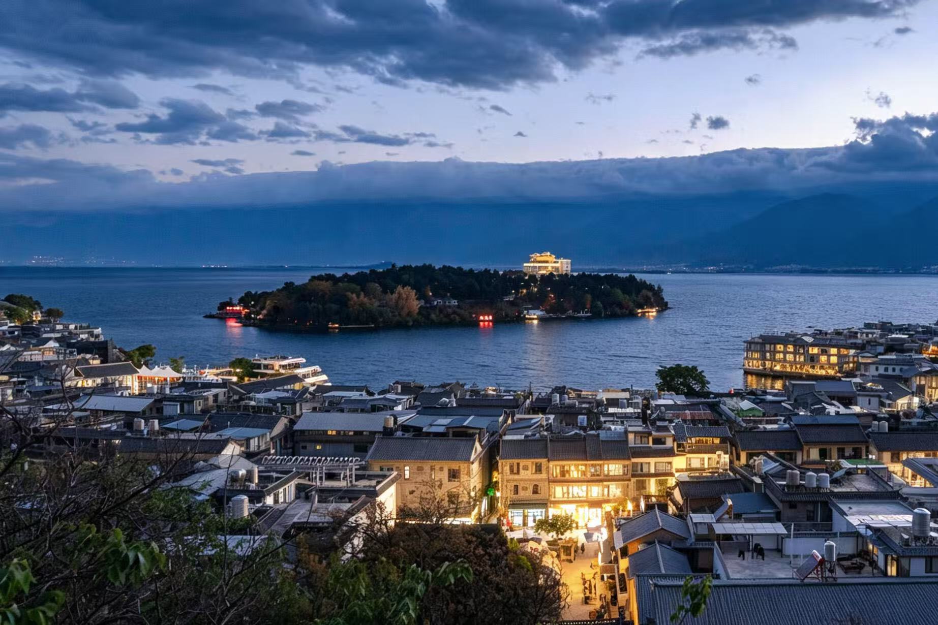 暮色降臨，雙廊古鎮燈火次第亮起。洱海上南詔風情島靜立，島上宮殿燈火璀璨。遠處蒼山隱於深藍夜色，湖面泛著微光，古村與湖山相映，靜謐又富有菸火氣息