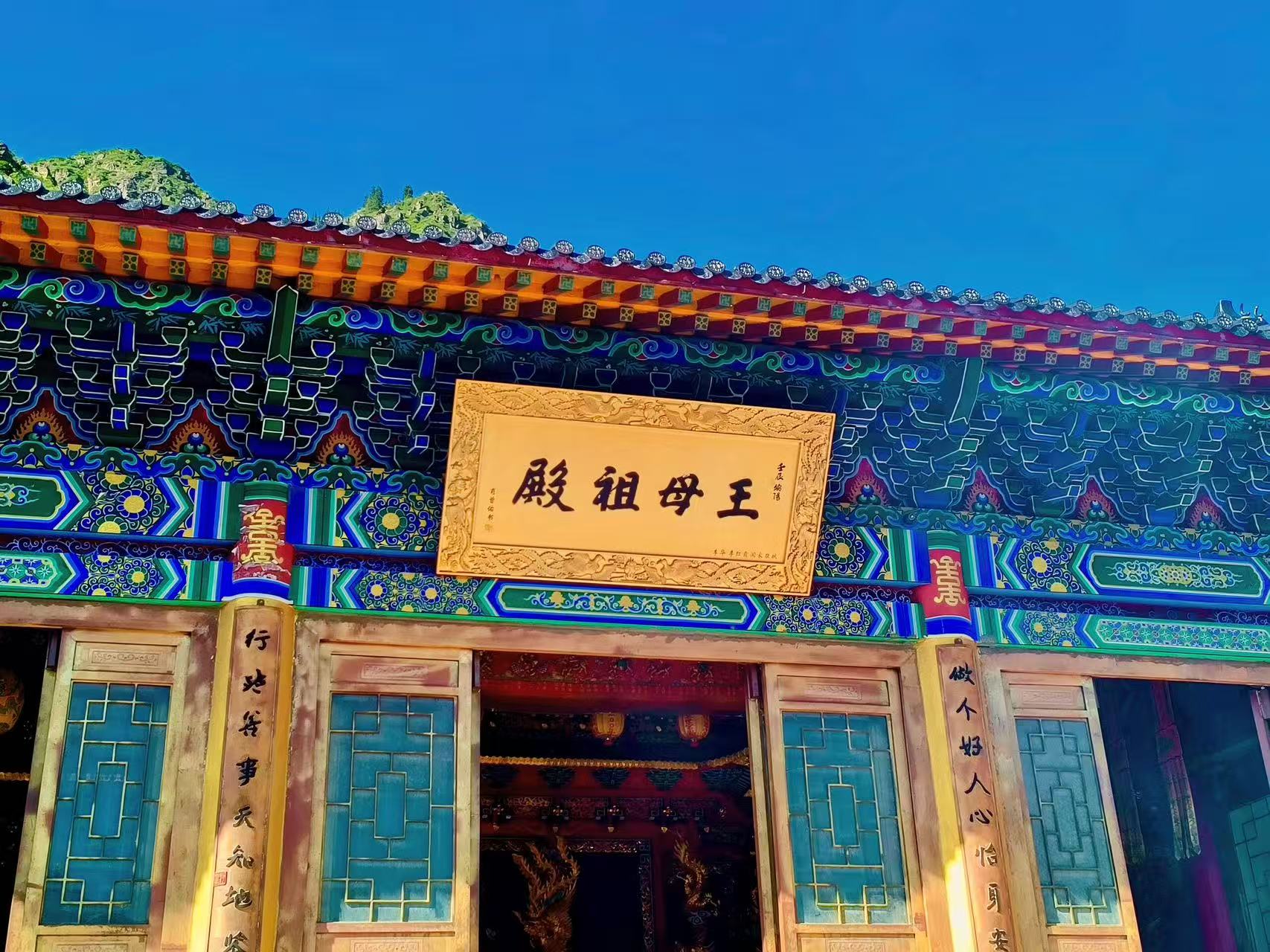中式古建王母祖殿，簷下彩畫繁複精美，藍綠紅黃雕飾層次豐富。正中懸掛金字牌匾，兩側木柱刻有楹聯，木格窗古樸雅緻，背後青山襯著晴空，古色古香，盡顯天池人文道觀古韻