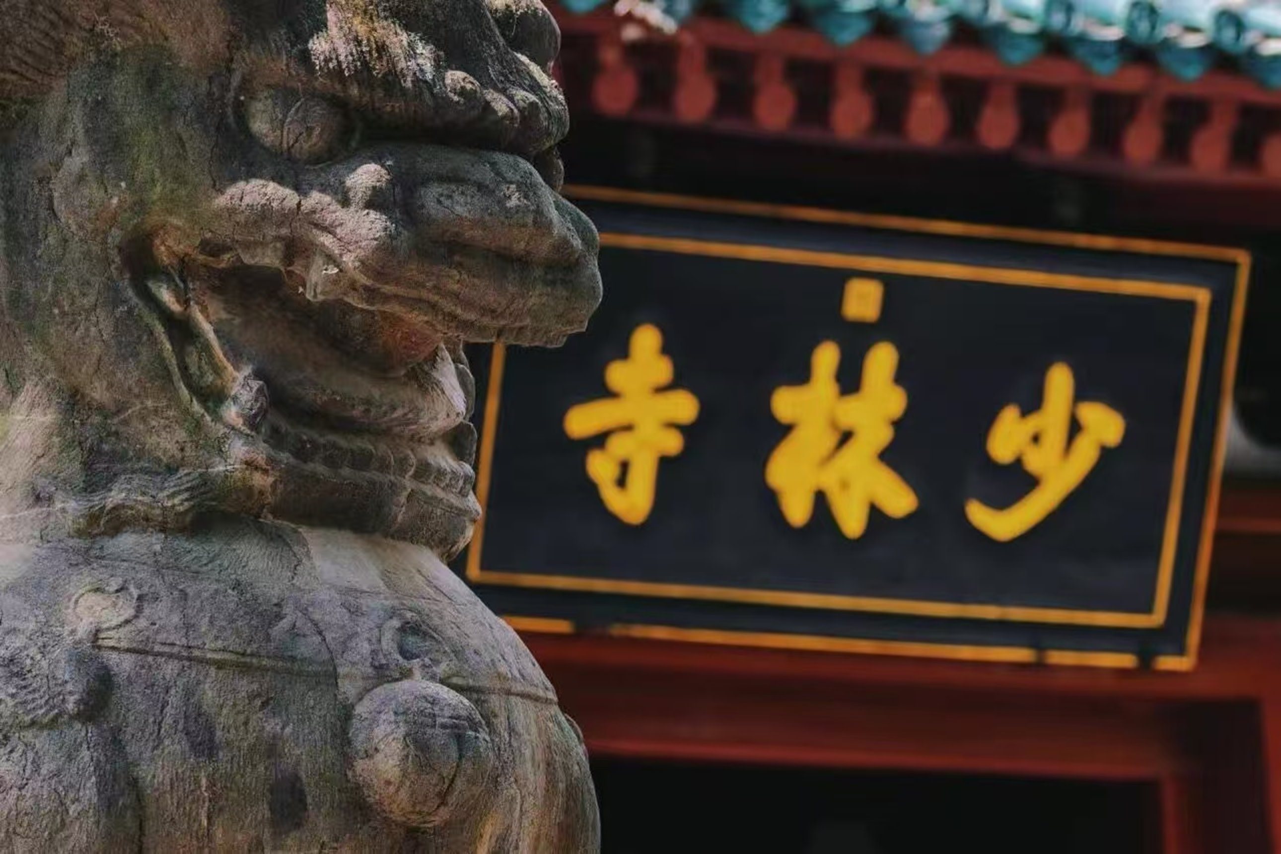 畫面主體是一尊飽經風霜的石獅子，紋理斑駁、神態威嚴，它側對著鏡頭，守護在少林寺山門旁。背景是黑底金字的 “少林寺” 匾額，搭配硃紅飛簷與青瓦，古樸厚重，盡顯千年古剎的莊重氣場