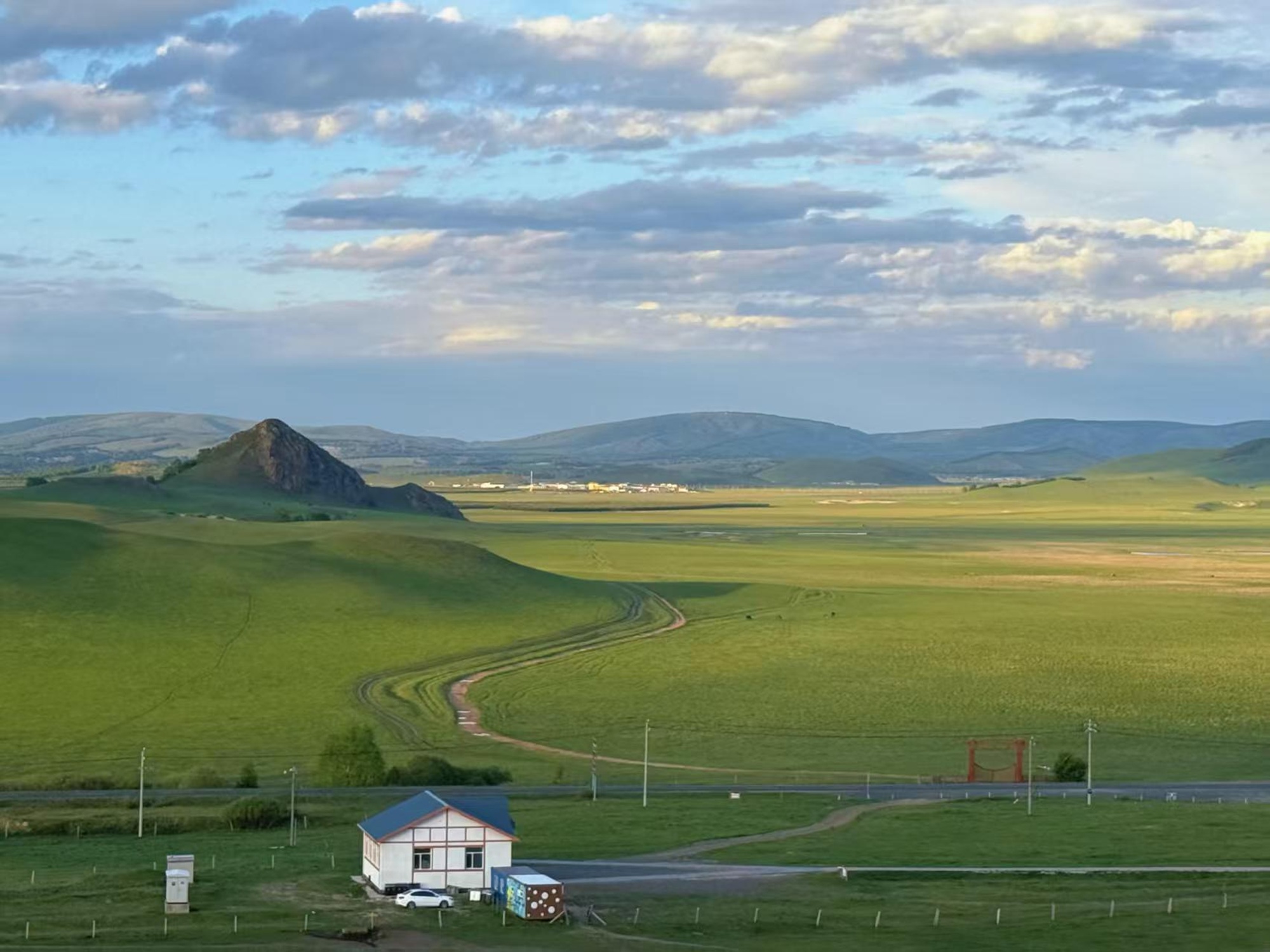 Across the vast grasslands, winding tire tracks mark the path of freedom, where a cabin stands in tranquility. Journey to this green expanse and encounter a poetic horizon.