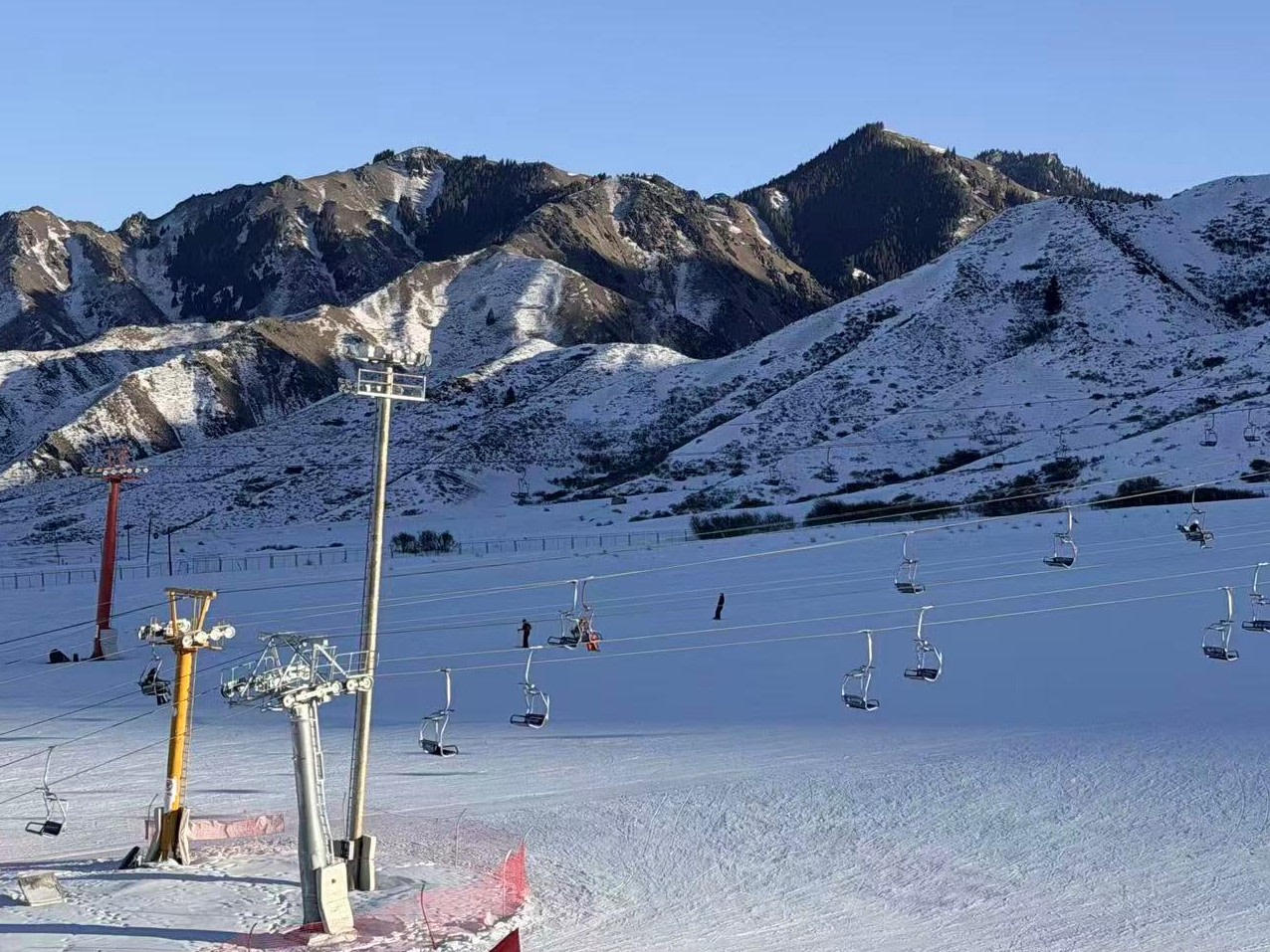 開闊雪場依山而建，連綿雪山錯落鋪展，明暗光影勾勒山體層次。多條吊椅纜車線路橫跨雪面，各色纜車支架矗立雪場，零星雪友分佈雪道，晴空萬里，山野雪景壯闊，盡顯雪場開闊的山地風貌