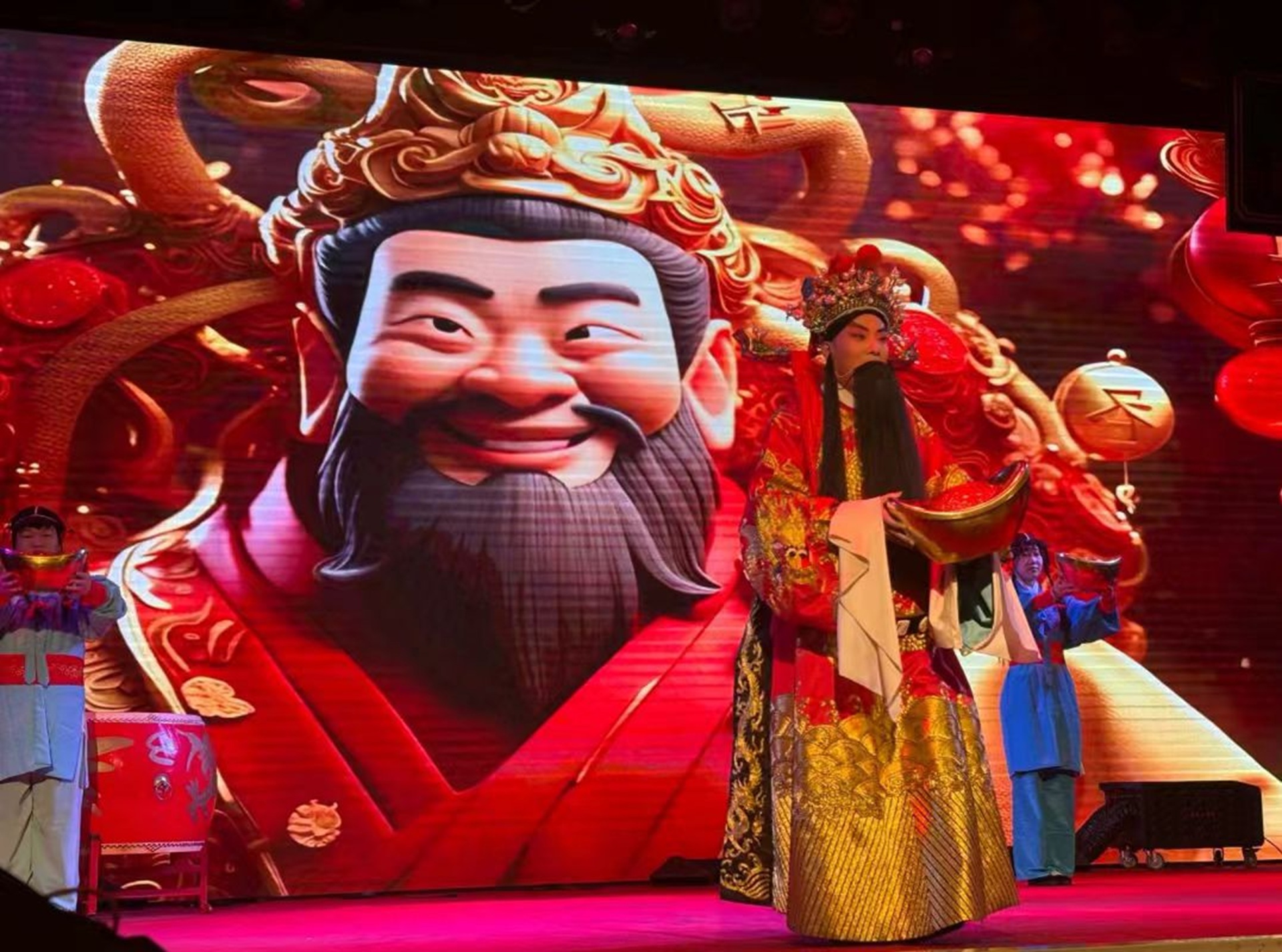 舞臺上演員扮財神，著紅黃戲服捧寶盆，背景大屏顯卡通財神像，氛圍喜慶紅火