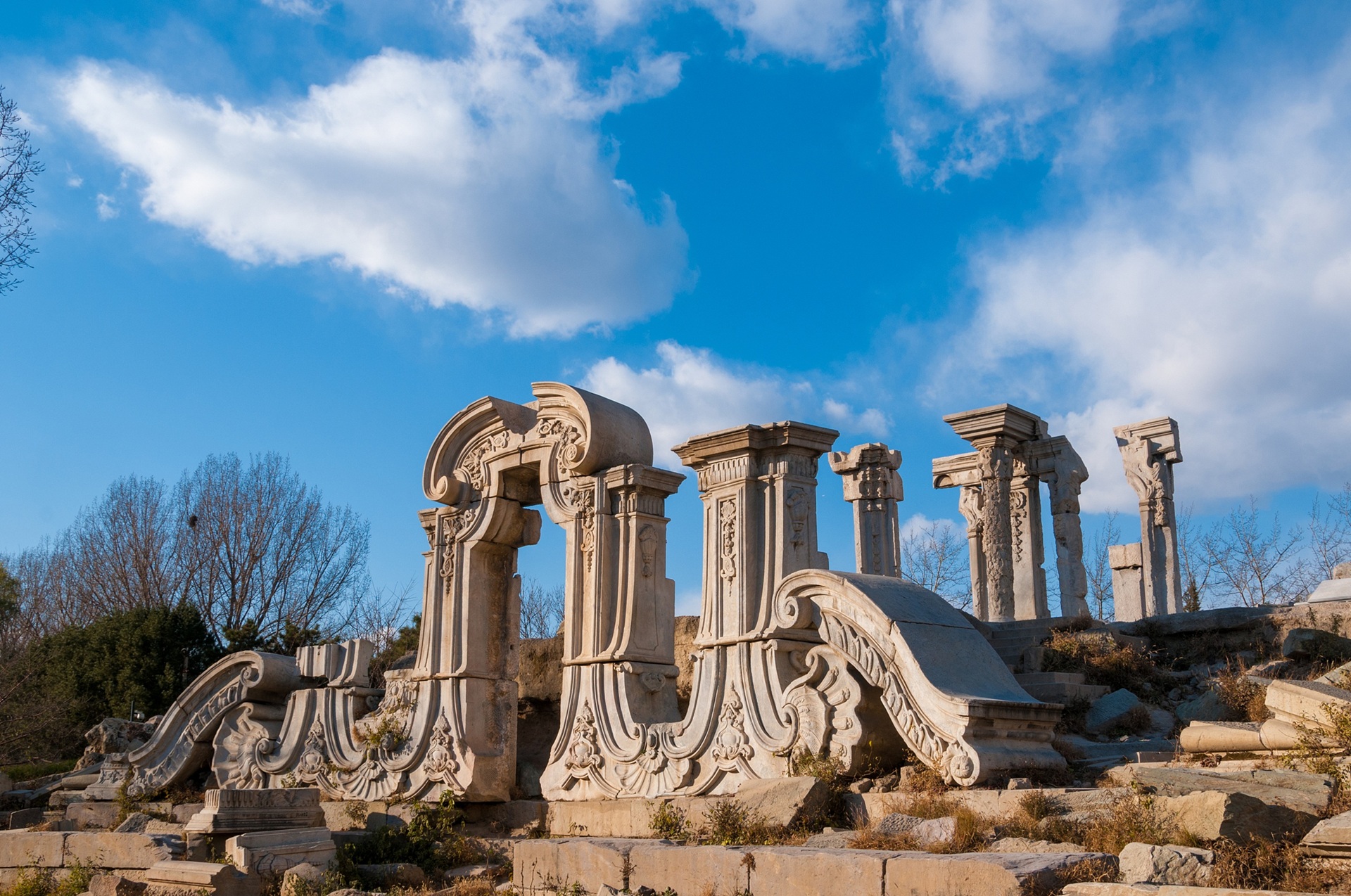 圓明園西洋樓遺址在晴空下靜默矗立，漢白玉雕花殘柱訴說著歷史滄桑，藍天白雲映襯下，盡顯文明的厚重與歷史的震撼