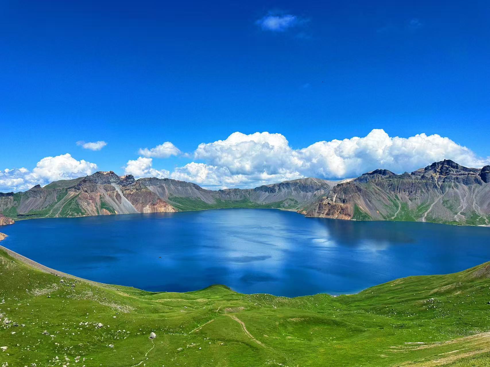 開闊視角完整展現高山天池，湖面如藍寶石般透亮，四周環繞層疊斑斕的火山岩壁，山麓鋪著綿軟翠綠草甸。澄澈藍天浮著大朵白雲，山水草甸相映，風光純淨遼闊，盡顯天池原生態絕美景緻