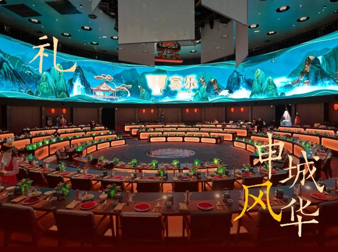 環形佈局的餐席層層排開，桌面擺著紅釉餐具與青綠裝飾器皿，暖光勾勒出宴席的雅緻氛圍；上方是 360 度環繞大屏，水墨山水、亭臺樓閣的國風畫面鋪滿整個視野，“禮”“宴樂”“申城風華” 的題字點綴其間，既顯唐風宴飲的儀式感，又融入了上海（申城）的地域文化標識。