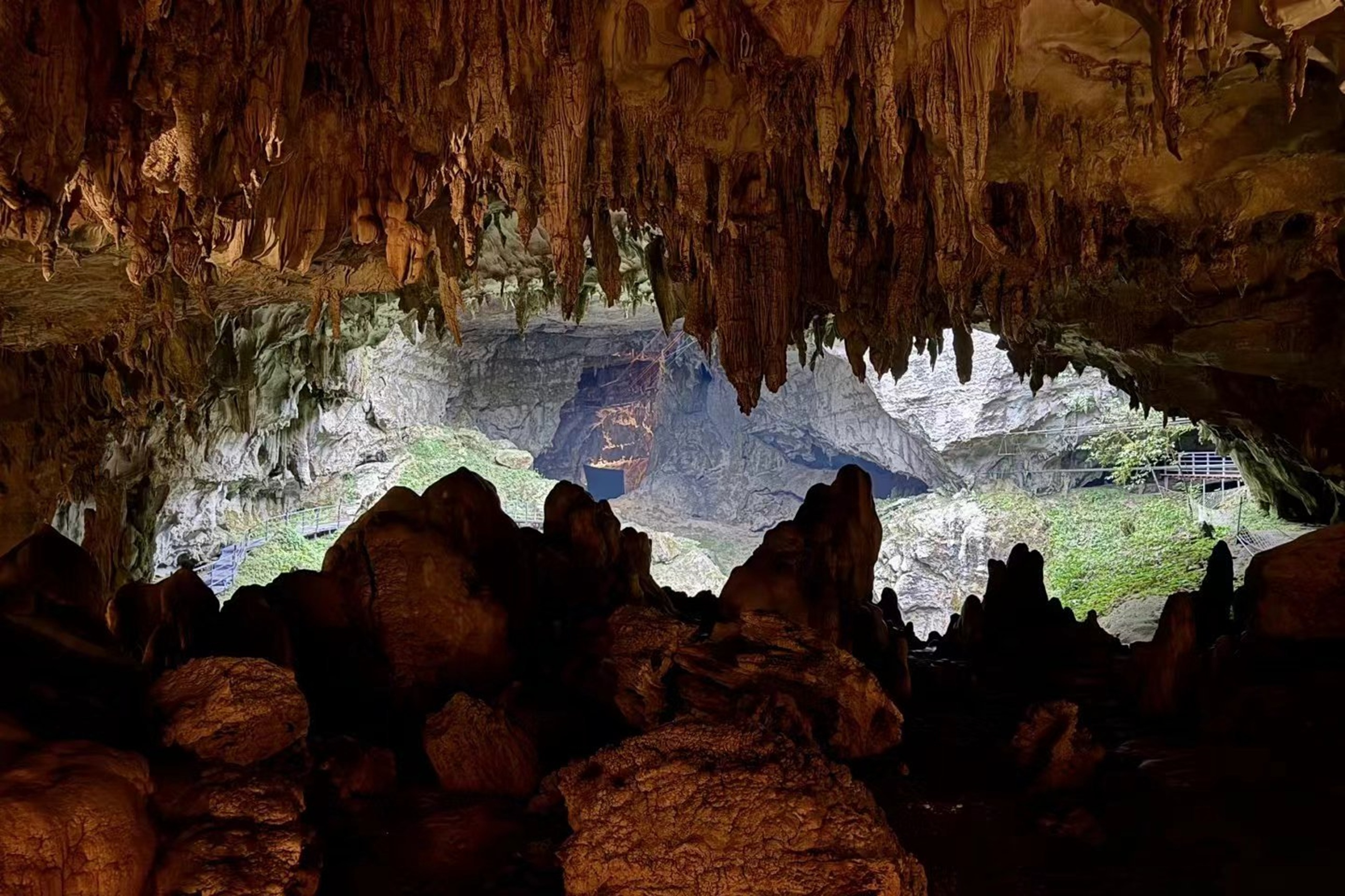 溶洞洞口鐘乳石如垂簾般掛滿穹頂,洞外綠意與洞內暖光交織,巖壁紋理粗獷,彷彿踏入時光凝固的喀斯特秘境 溶洞洞口鐘乳石如垂簾般掛滿穹頂,洞外綠意與洞內暖光交織,巖壁紋理粗獷,彷彿踏入時光凝固的喀斯特秘境