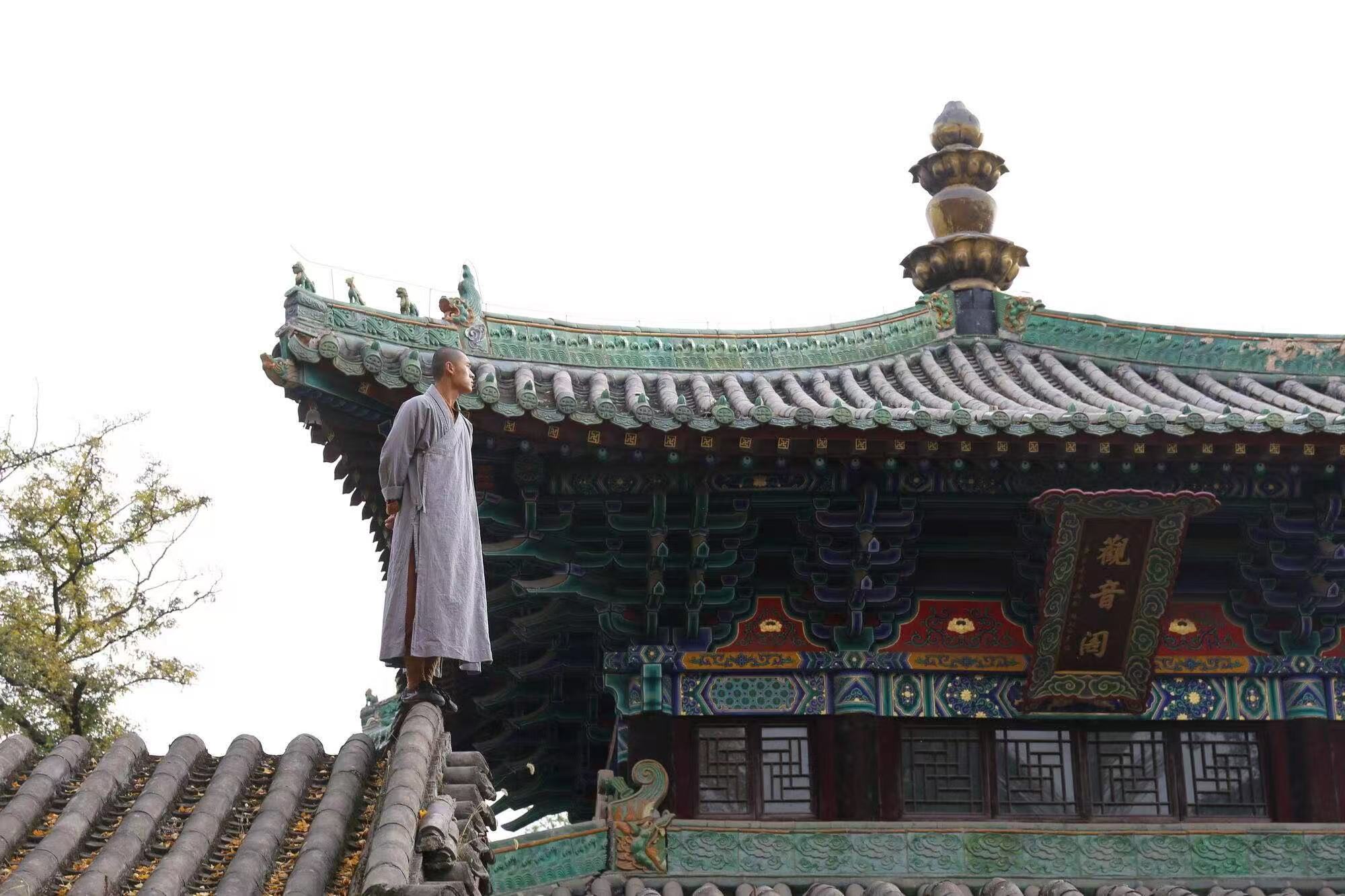 一位僧人立於觀音閣的飛簷屋脊之上，衣袂輕垂，背景是彩繪繁複的閣頂與鎏金塔剎。極簡的人物與華麗的古建形成視覺對比，既顯僧人清逸，也襯出觀音閣建築的精巧與莊重