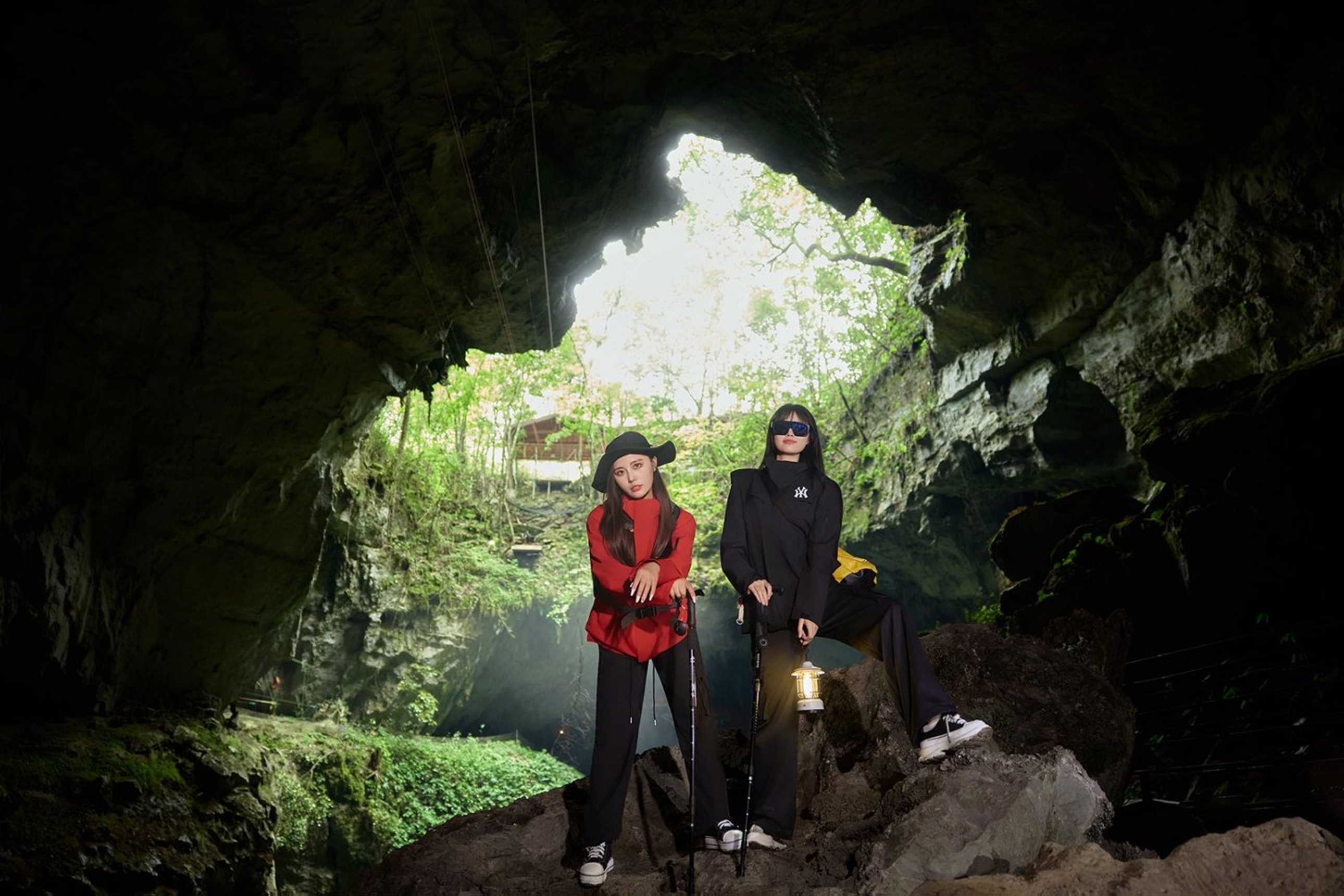 溶洞洞口處,兩位女孩立於明暗交界線,洞外綠意與洞內暗影交織,手持登山杖與煤油燈,盡顯探險前的期待與酷感 溶洞洞口處,兩位女孩立於明暗交界線,洞外綠意與洞內暗影交織,手持登山杖與煤油燈,盡顯探險前的期待與酷感