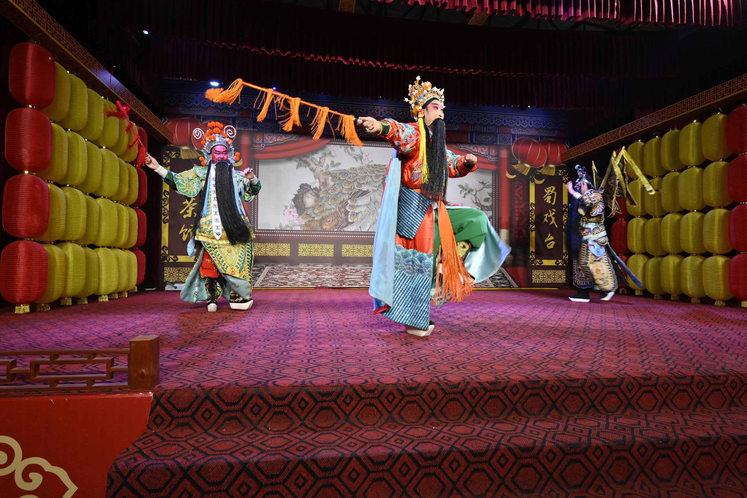 在掛滿紅黃燈籠的舞臺上表演。一位演員手持長鞭，動作矯健，另外兩位演員或立或動，傳統的舞臺佈景與演員的精彩演繹，展現出蜀戲的獨特風采