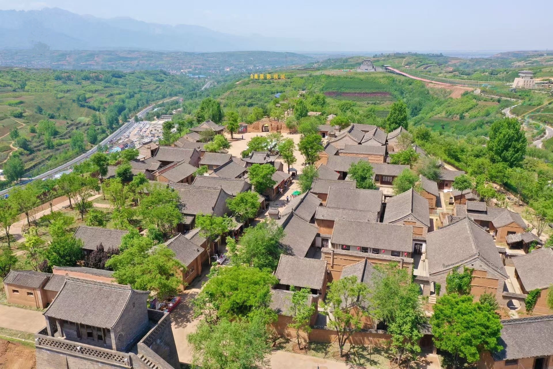 影視城佈局,古建群落錯落山間,綠樹掩映,遠處山巒、道路延伸,展現其依山而建的格局,凸顯歷史建築群與自然環境的融合之美 影視城佈局,古建群落錯落山間,綠樹掩映,遠處山巒、道路延伸,展現其依山而建的格局,凸顯歷史建築群與自然環境的融合之美