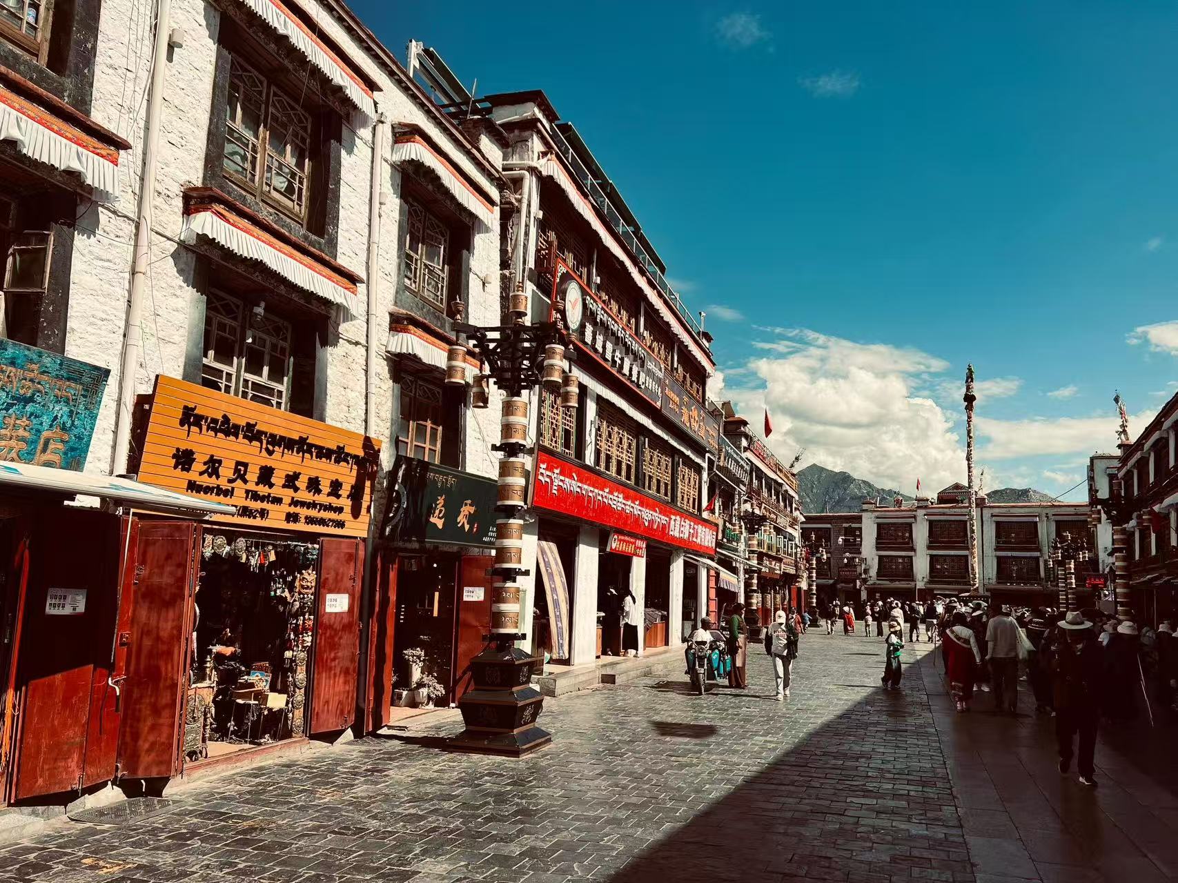 八廓街藏式商鋪沿街排布,白牆紅木門搭配雙語招牌,復古路燈立在石板路上。行人悠閒漫步,遠處可見山巒,湛藍天空襯著藏式建築,菸火與藏地人文風情融為一體 八廓街藏式商鋪沿街排布,白牆紅木門搭配雙語招牌,復古路燈立在石板路上。行人悠閒漫步,遠處可見山巒,湛藍天空襯著藏式建築,菸火與藏地人文風情融為一體