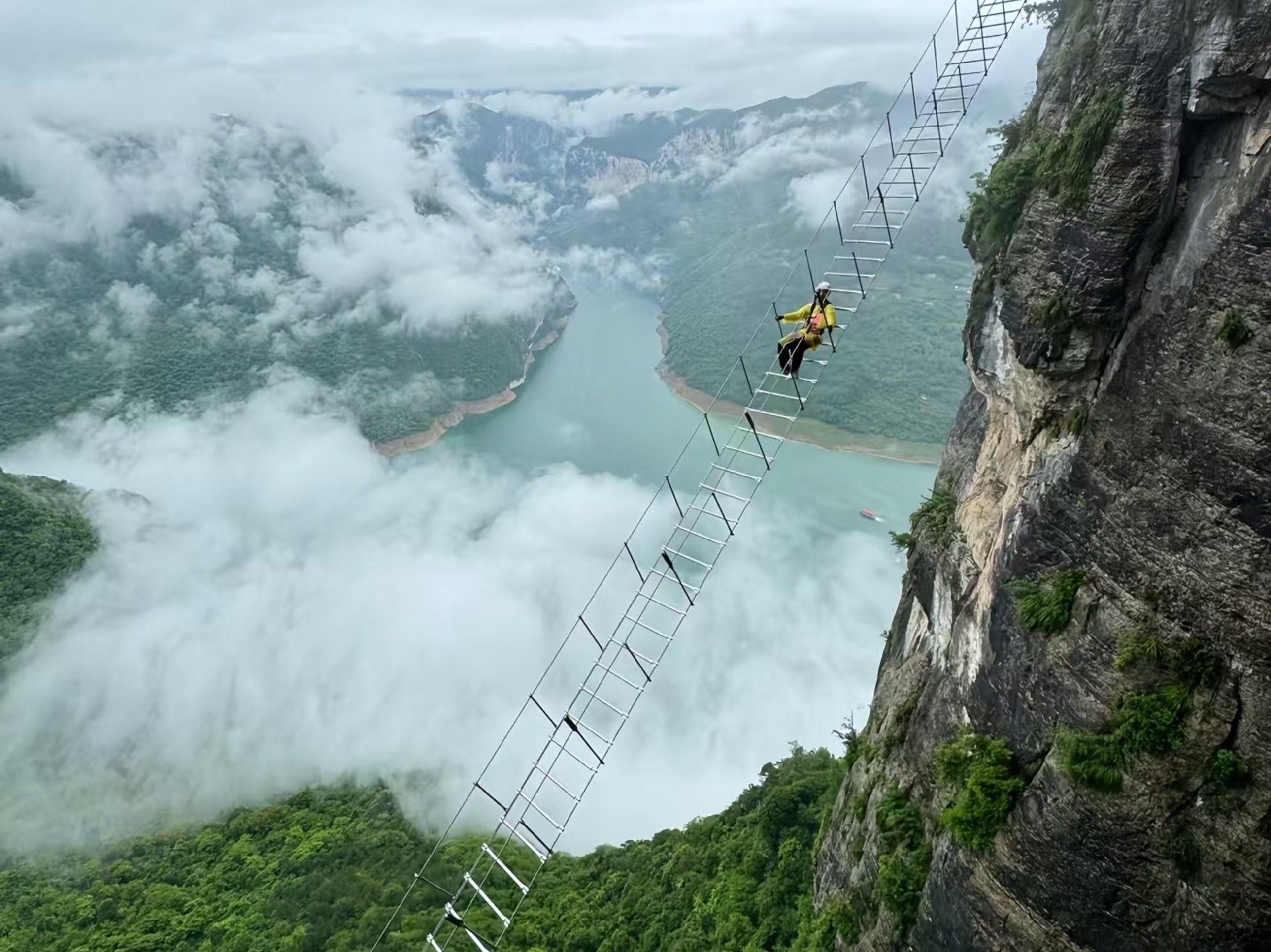 穿黃衣的人在雲霧繚繞的高空梯上前行，江面隱於霧中，崖壁冷峻，朦朧裡透著高空探險的神秘與膽魄