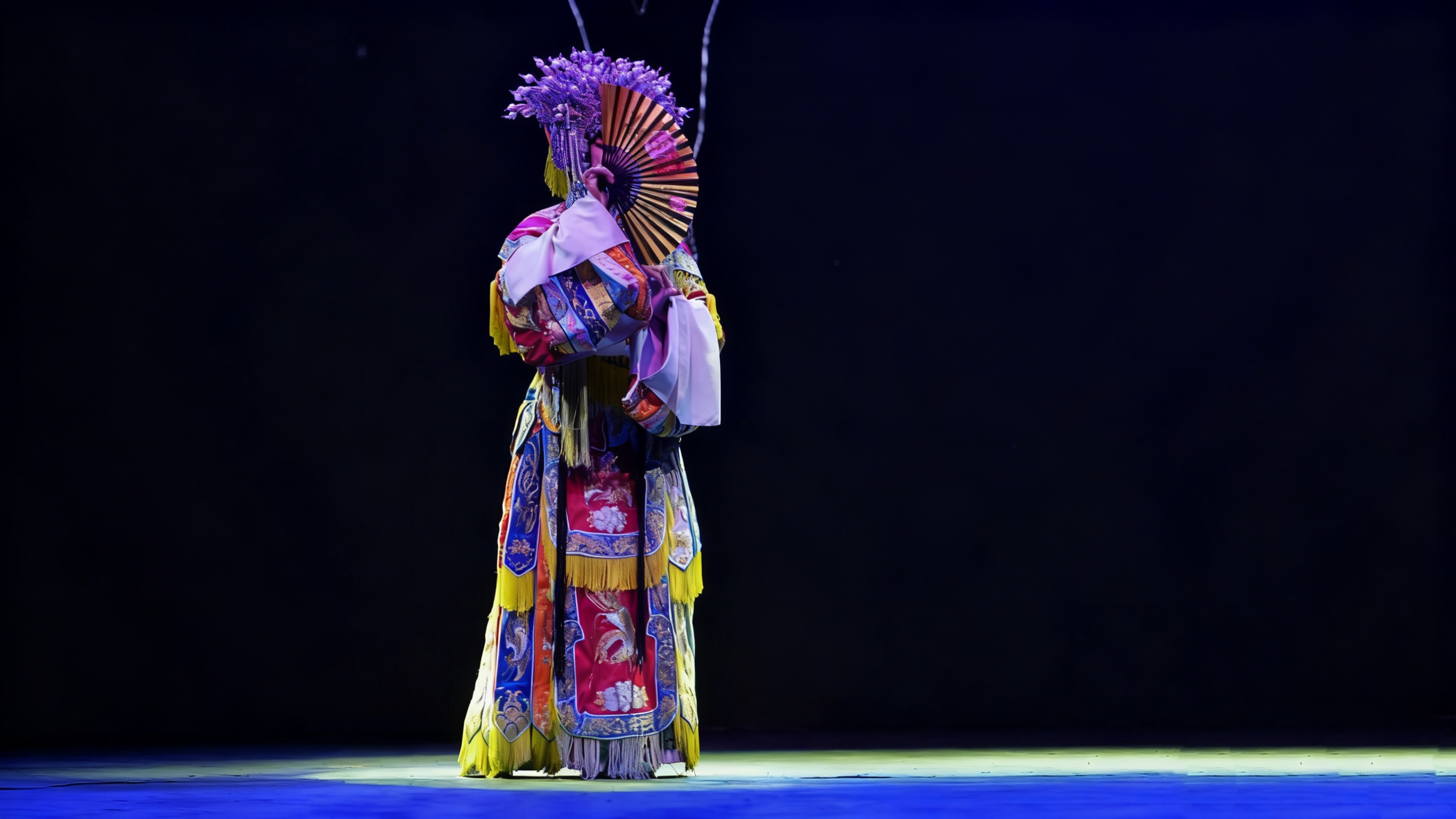 京劇旦角演員立於舞臺中央,頭戴紫色的鳳冠,手持一把精緻的摺扇。流蘇隨著動作輕輕搖曳,在深色背景的襯托下,服飾的色彩與紋樣格外奪目 京劇旦角演員立於舞臺中央,頭戴紫色的鳳冠,手持一把精緻的摺扇。流蘇隨著動作輕輕搖曳,在深色背景的襯托下,服飾的色彩與紋樣格外奪目