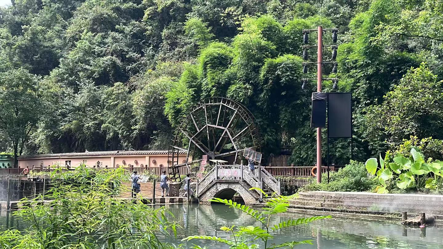 清澈的溪流邊，一座巨大的木質水車靜靜佇立，旁邊的石拱橋與步道上的行人相映成趣
