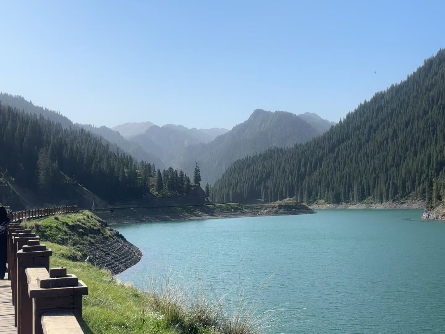木質觀景棧道沿湖岸延伸，一側是覆著青草的坡地，另一側湖水呈通透青碧。峽谷兩側長滿密集雲杉，層層遠山薄霧輕籠，湖面開闊靜謐，山野幽深大氣，風光遼闊舒展