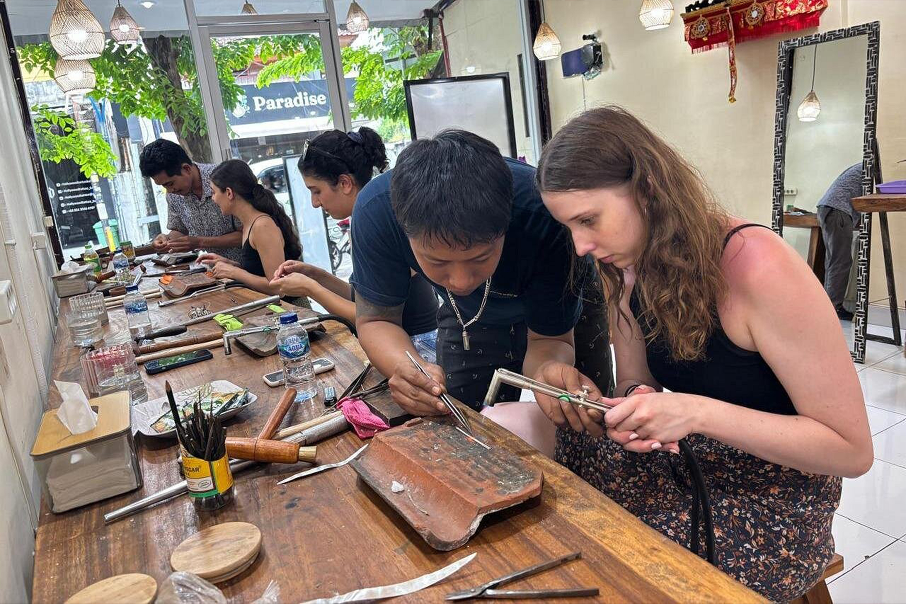 An instructor guides workshop participants in making silver jewelry using soldering techniques at a work table.