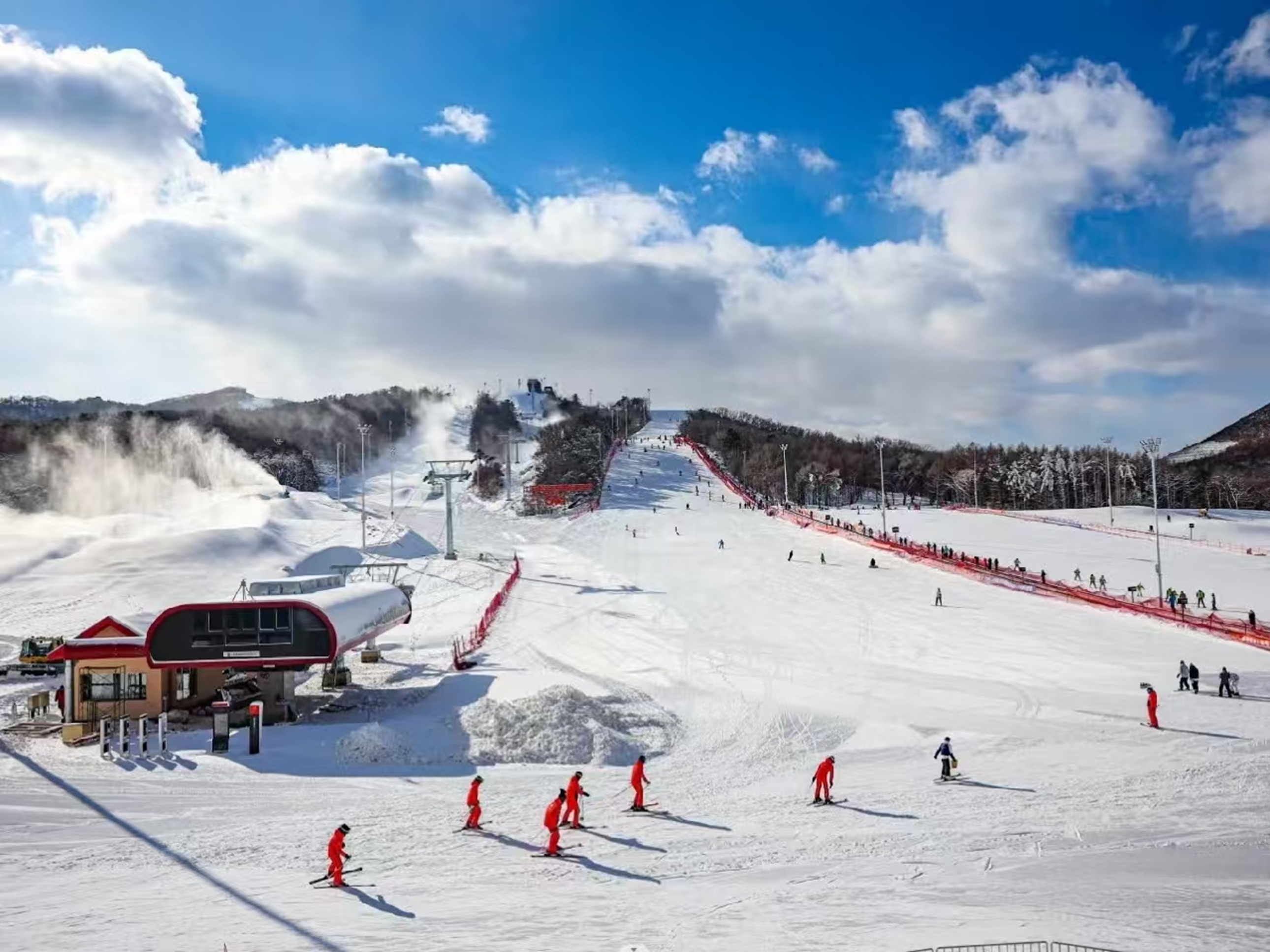 開闊雪場裡，造雪機噴雪霧，紅衣滑雪者成排滑行，纜車與建築錯落，晴空襯得雪場明媚又熱鬧