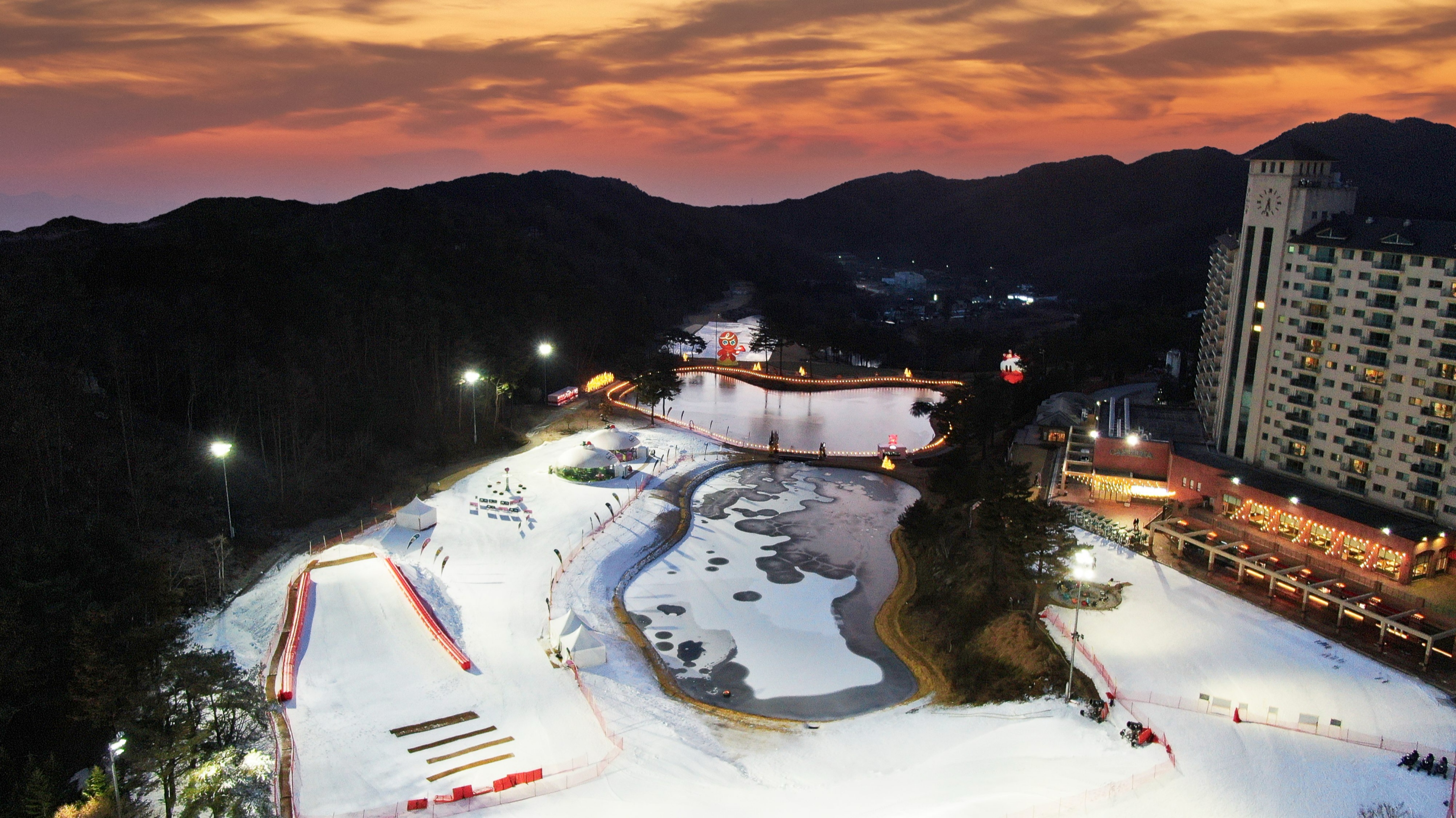 橡樹谷黃昏:在韓國雙板滑雪度假村的日落餘暉下,公寓燈火通明,池塘結冰,人們在雪中嬉戲。 橡樹谷黃昏:在韓國雙板滑雪度假村的日落餘暉下,公寓燈火通明,池塘結冰,人們在雪中嬉戲。