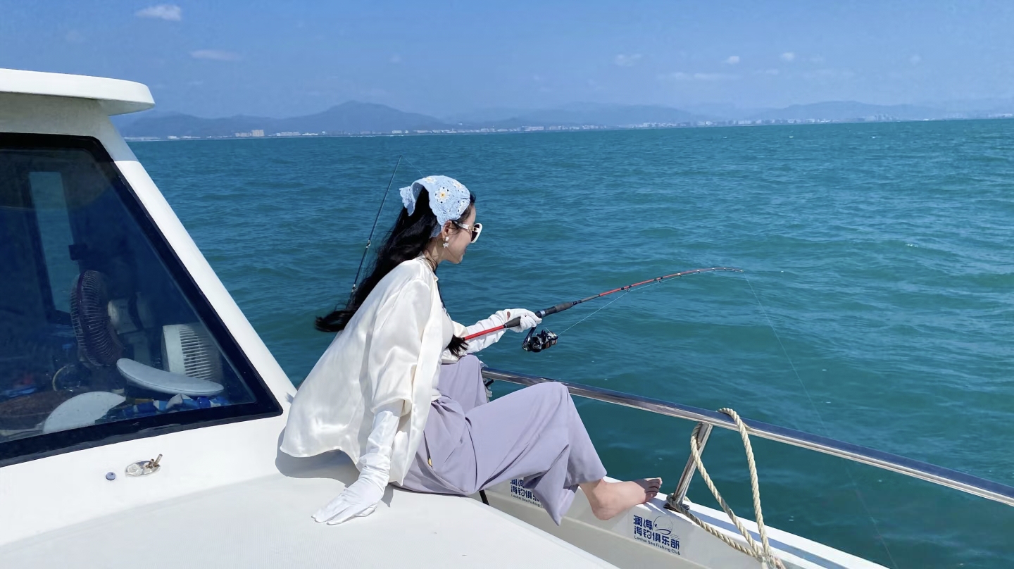 A blue headscarf sets off a fair neck, a surfcasting rod leans against the railing, the fishing line is cast into the distance, and the skirt and headscarf sway gently in the breeze.