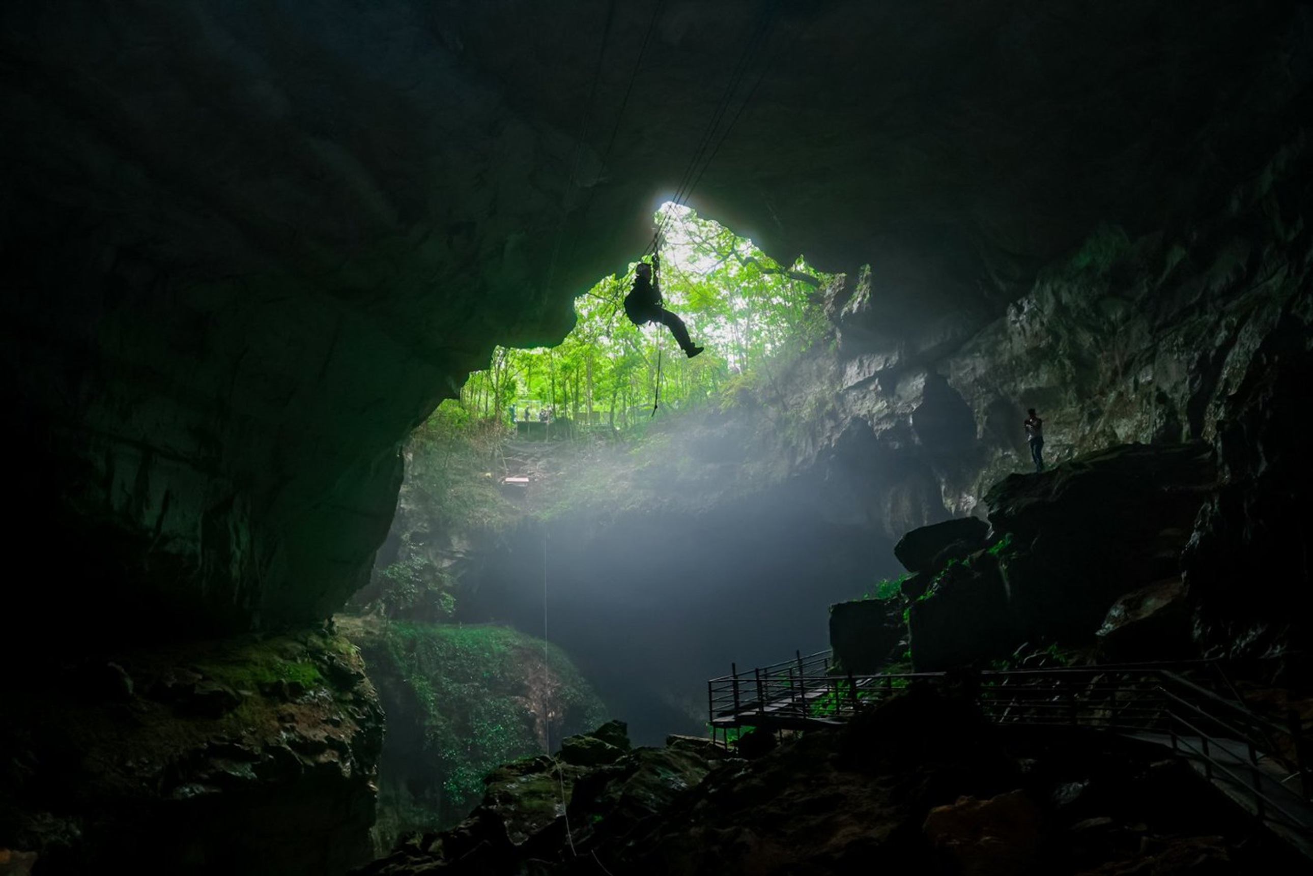 探險者乘溜索穿越溶洞天坑,洞口綠意與洞內暗影交織,光束穿透薄霧,彷彿闖入地心與森林的交界秘境,刺激又治癒 探險者乘溜索穿越溶洞天坑,洞口綠意與洞內暗影交織,光束穿透薄霧,彷彿闖入地心與森林的交界秘境,刺激又治癒