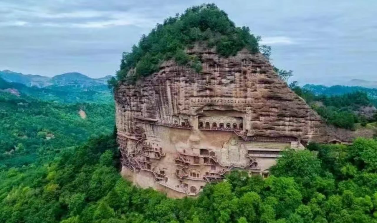 麥積山石窟位於甘肅省天水市，是中國四大石窟之一。畫面中，這座形似麥垛的孤峰拔地而起，山體表面密佈著層層疊疊的石窟群，棧道如游龍般蜿蜒纏繞崖壁，連接著數百個洞窟，展現出“峭壁之間鐫石成佛”的奇觀。石窟始鑿於北魏，以精美的泥塑藝術聞名於世，被譽為“東方雕塑館”。山體紅巖與周邊蒼翠的森林形成鮮明對比，自然景觀與人文遺產完美融合。作為世界文化遺產，麥積山不僅承載著千年佛教文化，更以獨特的建築智慧與藝術價值，成為絲綢之路上璀璨的文化地標。