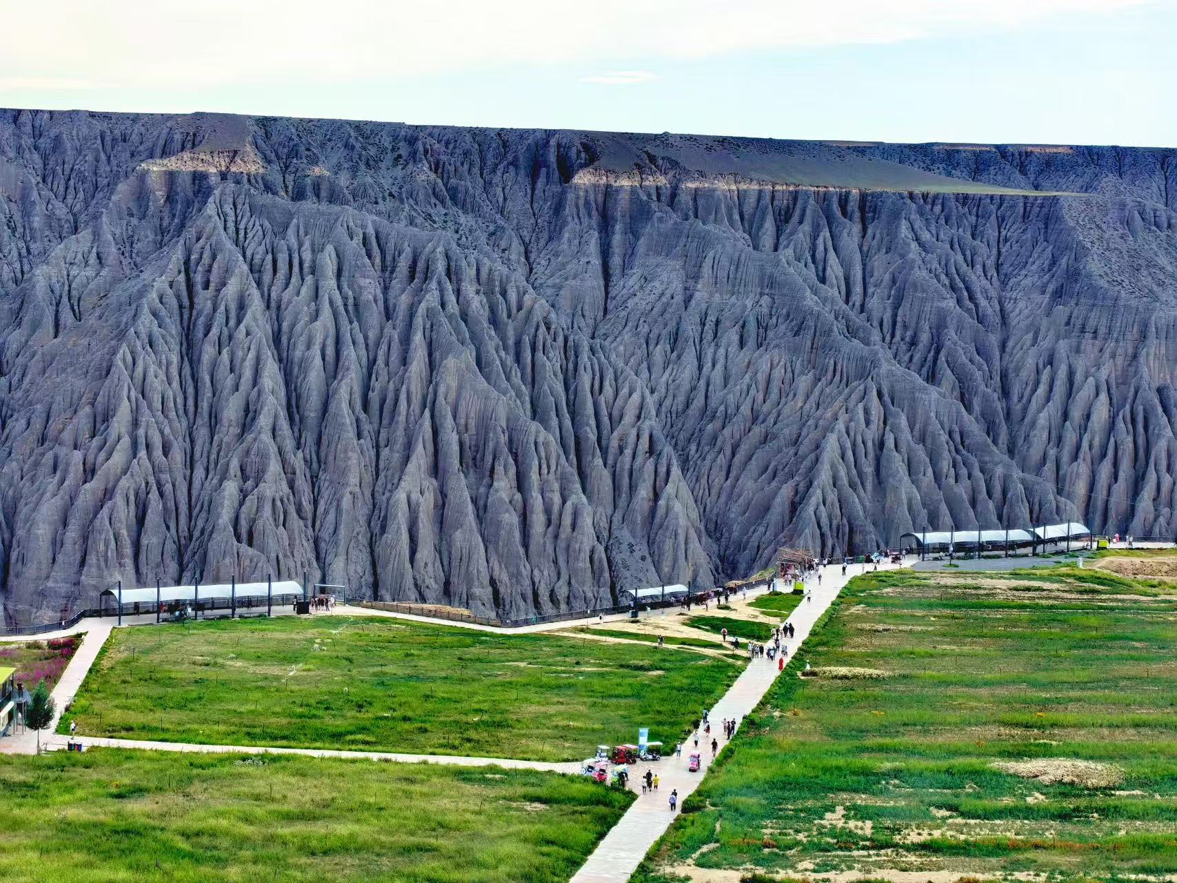 近距離直面峽谷崖壁，千萬年流水侵蝕出密集褶皺，灰黑色巖壁層層堆疊。崖前開闊青草地鋪展，步道上游客緩緩行走，配套休憩長廊坐落一旁，剛硬地貌搭配柔軟草甸，剛柔對比極具視覺衝擊