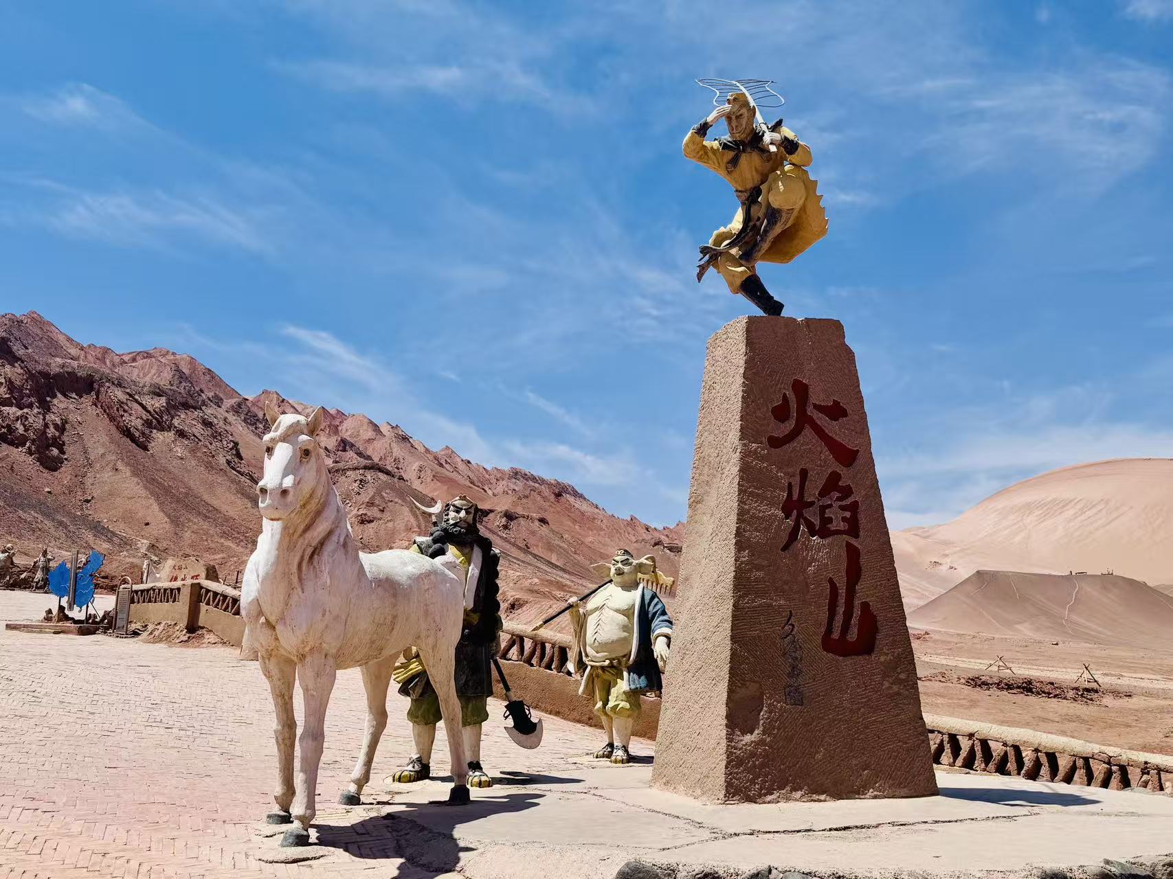 火焰山景區立石碑刻紅字，碑頂塑孫悟空遠眺雕像，一旁白龍馬、八戒、沙僧塑像完整復刻西遊師徒。赤紅荒山沙丘作背景，借神話 IP 凸顯景區西遊文化特色