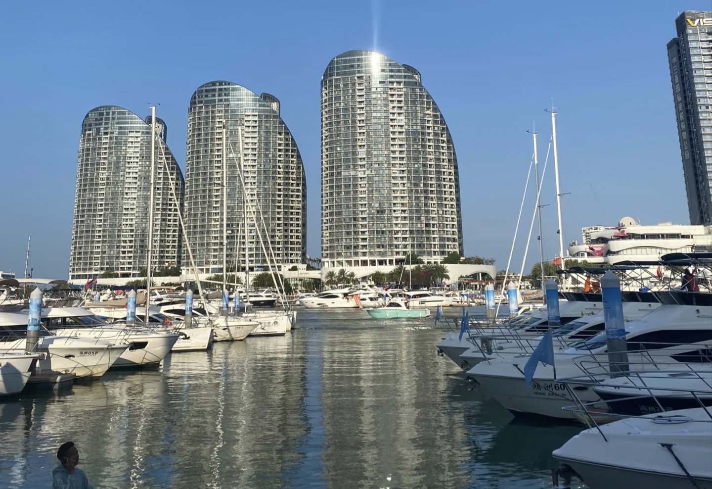 The sunlight splashed bright, shimmering spots of light across the building. The dock was filled with white yachts, their masts reaching diagonally towards the sky, and the water reflected the hulls and buildings.