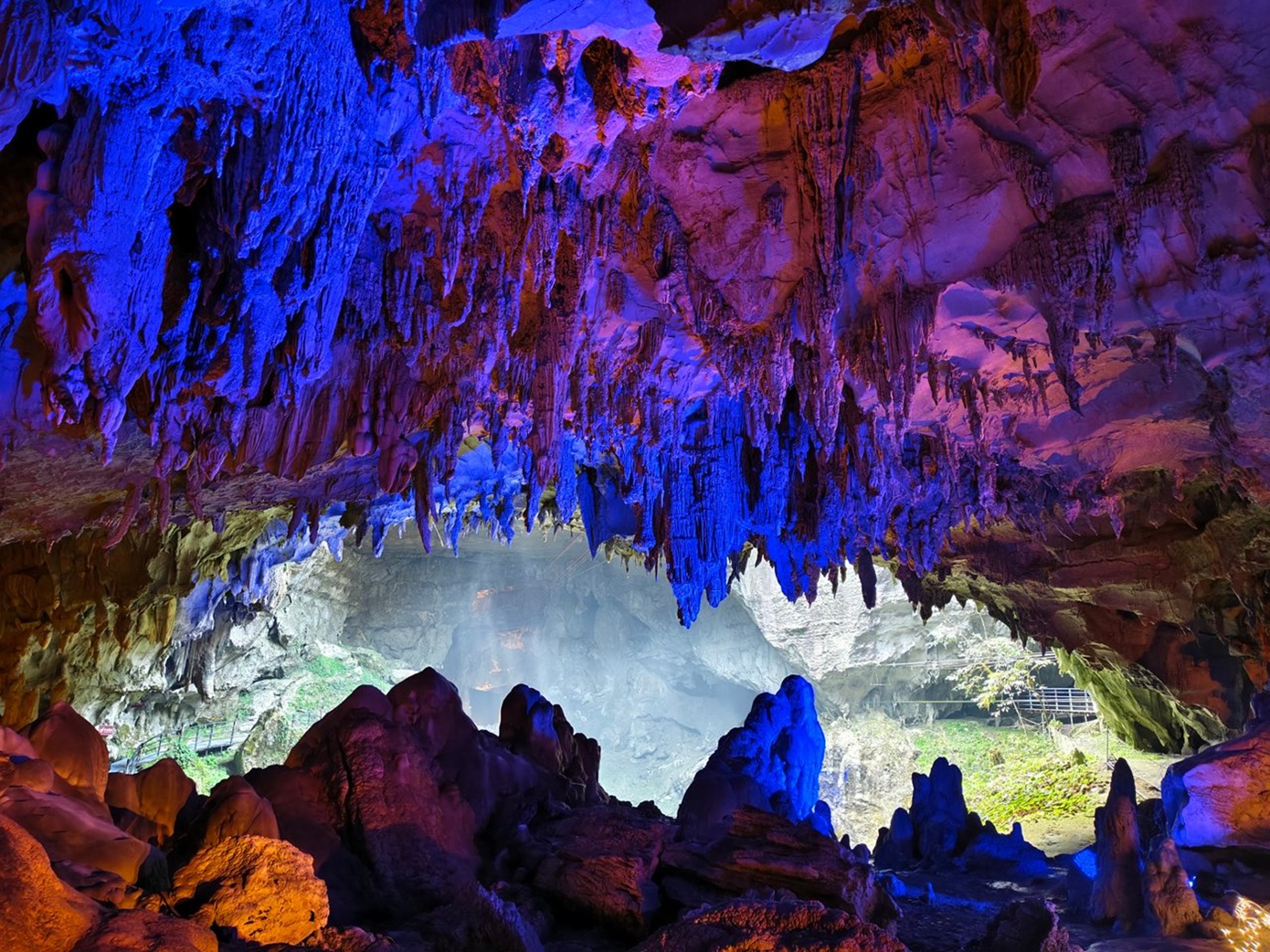 溶洞洞口被藍紫燈光點亮,鐘乳石如冰稜垂掛,洞外綠意隱約可見,光影交織成夢幻的喀斯特地貌奇觀 溶洞洞口被藍紫燈光點亮,鐘乳石如冰稜垂掛,洞外綠意隱約可見,光影交織成夢幻的喀斯特地貌奇觀