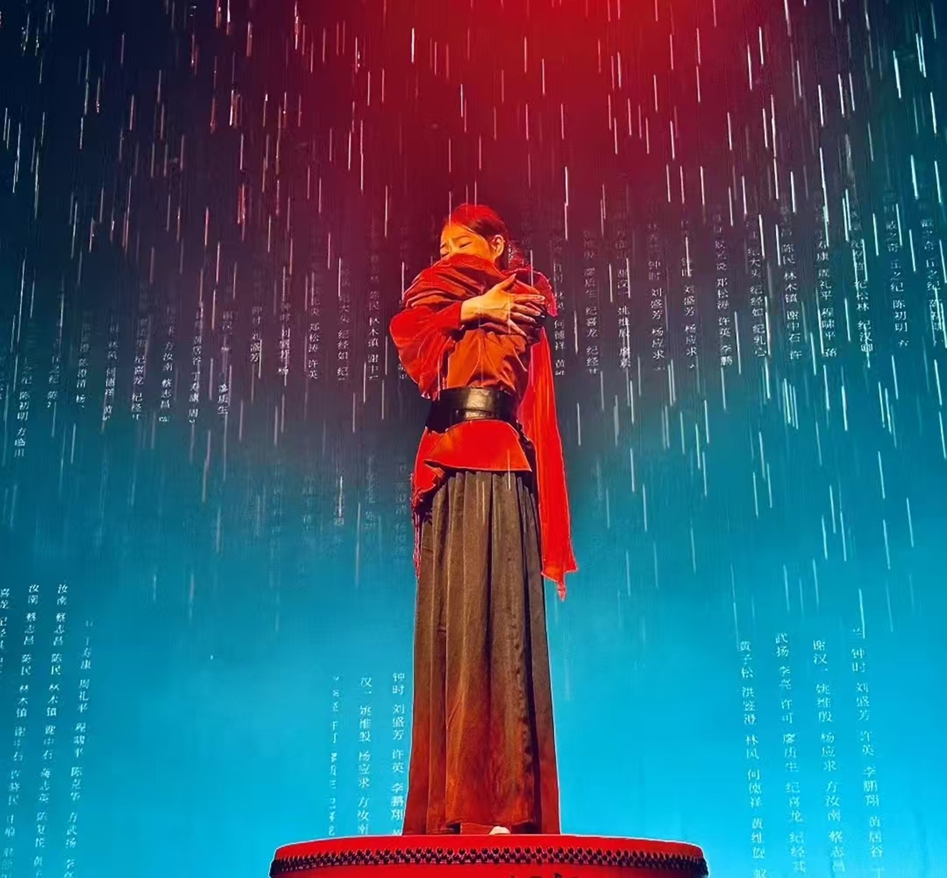 紅衣演員立於鼓上抱臂，背景雨幕襯著人名文字，紅藍光影裡滿是故事感