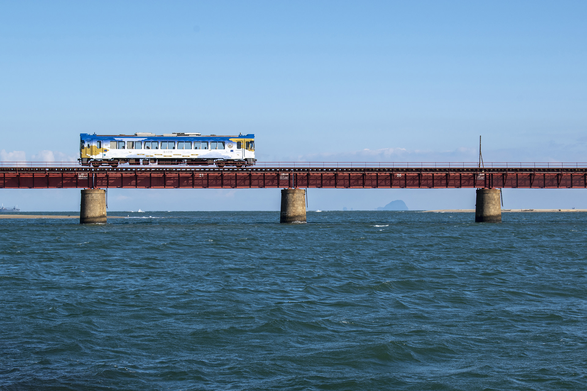 京都丹後海上列車沿著日本海岸線緩緩行駛，窗外展開的是一幅連綿不絕的海岸風景畫。湛藍的海水隨著光線變化呈現不同層次的藍色，遠方山巒層疊起伏，與海天交界相互映襯。列車穿越小漁村與靜謐港灣，傳統木造建築與停泊的漁船點綴其間，構成一種寧靜而富有生活氣息的沿海景致。