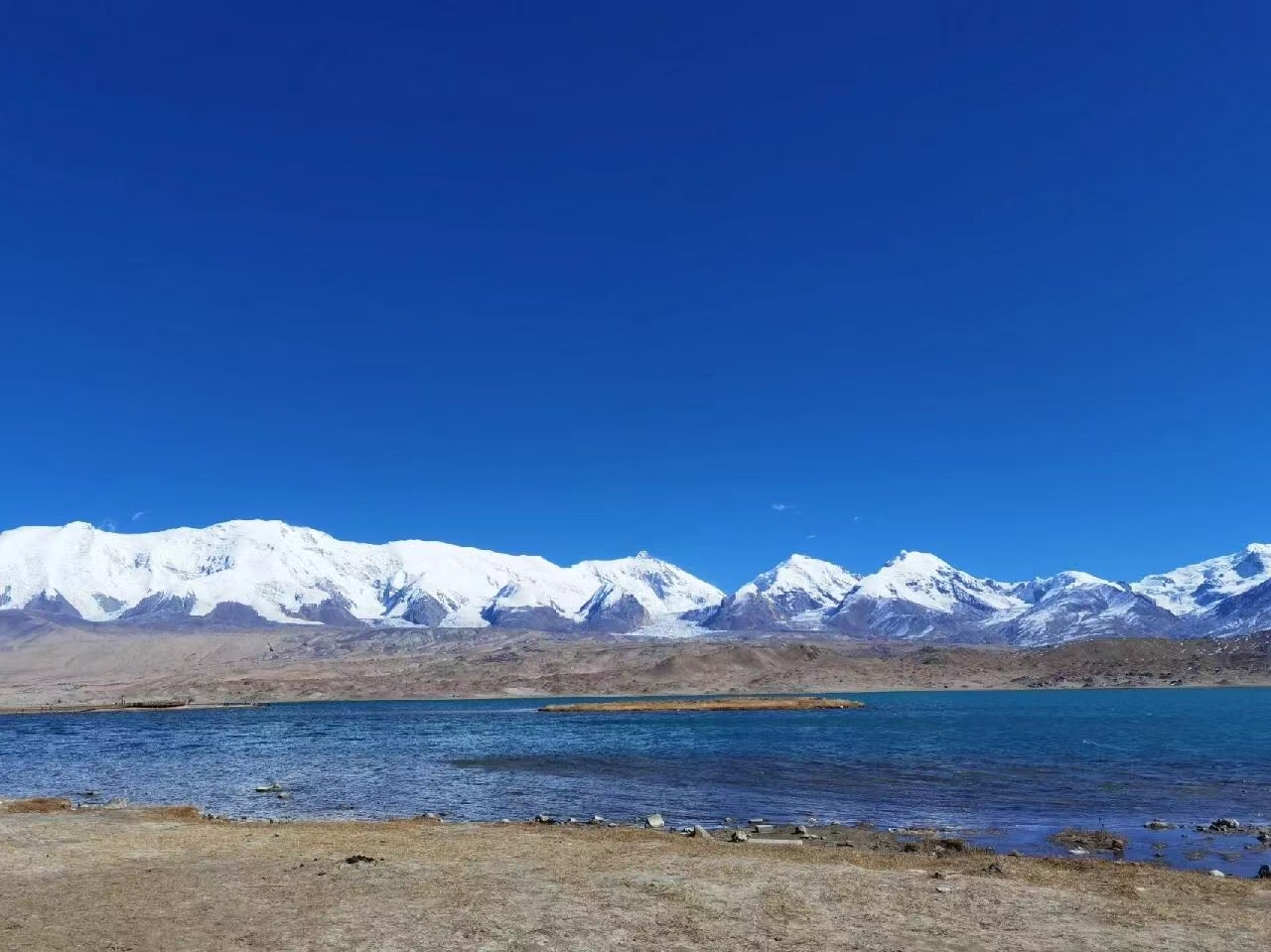 湛藍的天空下，慕士塔格峰與連綿的雪山群峰巍峨聳立，峰頂的白雪在陽光下熠熠生輝。山腳下的湖泊泛著幽藍的波光，展現出帕米爾高原的極致純淨