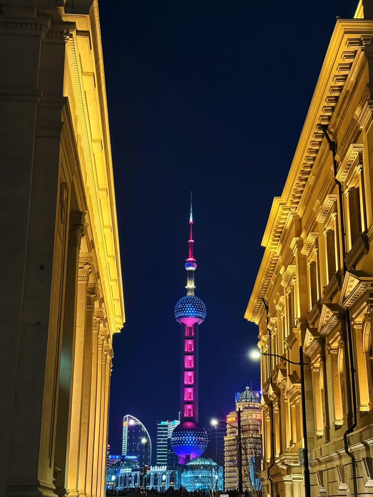 外灘街頭視角，兩側歐式古典建築的金色廊柱勾勒出絕佳框景。夜幕下，東方明珠塔以粉藍漸變燈光點亮天際，與建築輪廓相映，古典與現代碰撞，盡顯上海的包容與繁華