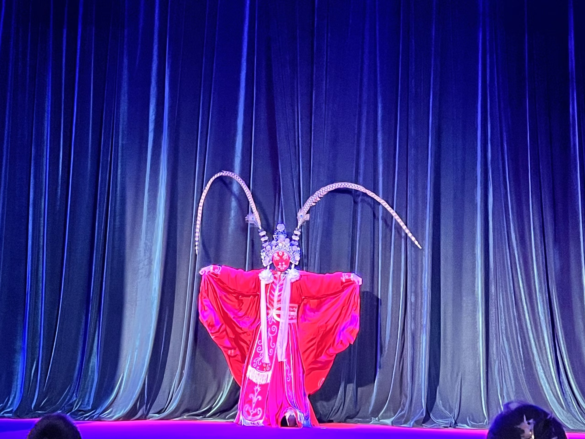 戲曲演員穿豔紅戲服，飾銀角頭飾，展袖立於藍幕前，妝容精緻，盡顯傳統戲曲的華彩