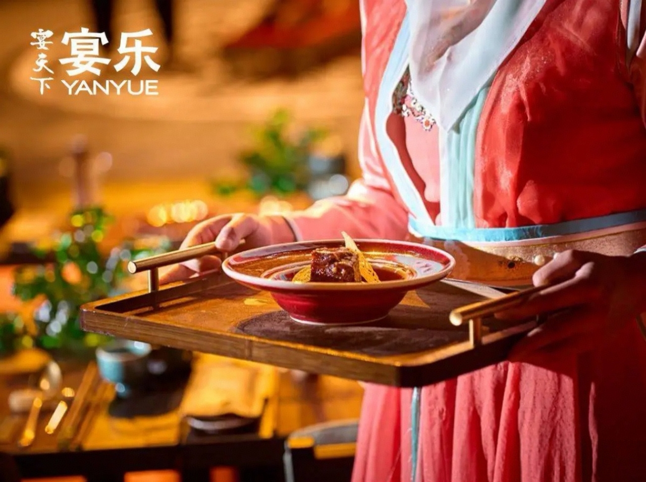 身著橙紅唐風襦裙（鑲淺藍邊飾，領口綴精緻紋樣）的侍女，手託木金相間的古式托盤，盤中是紅釉碗盛著的餐食（色澤濃郁的主菜配點綴食材）。暖黃燈光裹著餐具與服飾，背景隱約可見席間的青綠器皿與朦朧光暈，“宴天下 宴樂” 的標識襯出場景 —— 連上菜都成了古風儀式的一部分，讓食客從菜品端出的瞬間就浸入唐宴氛圍。