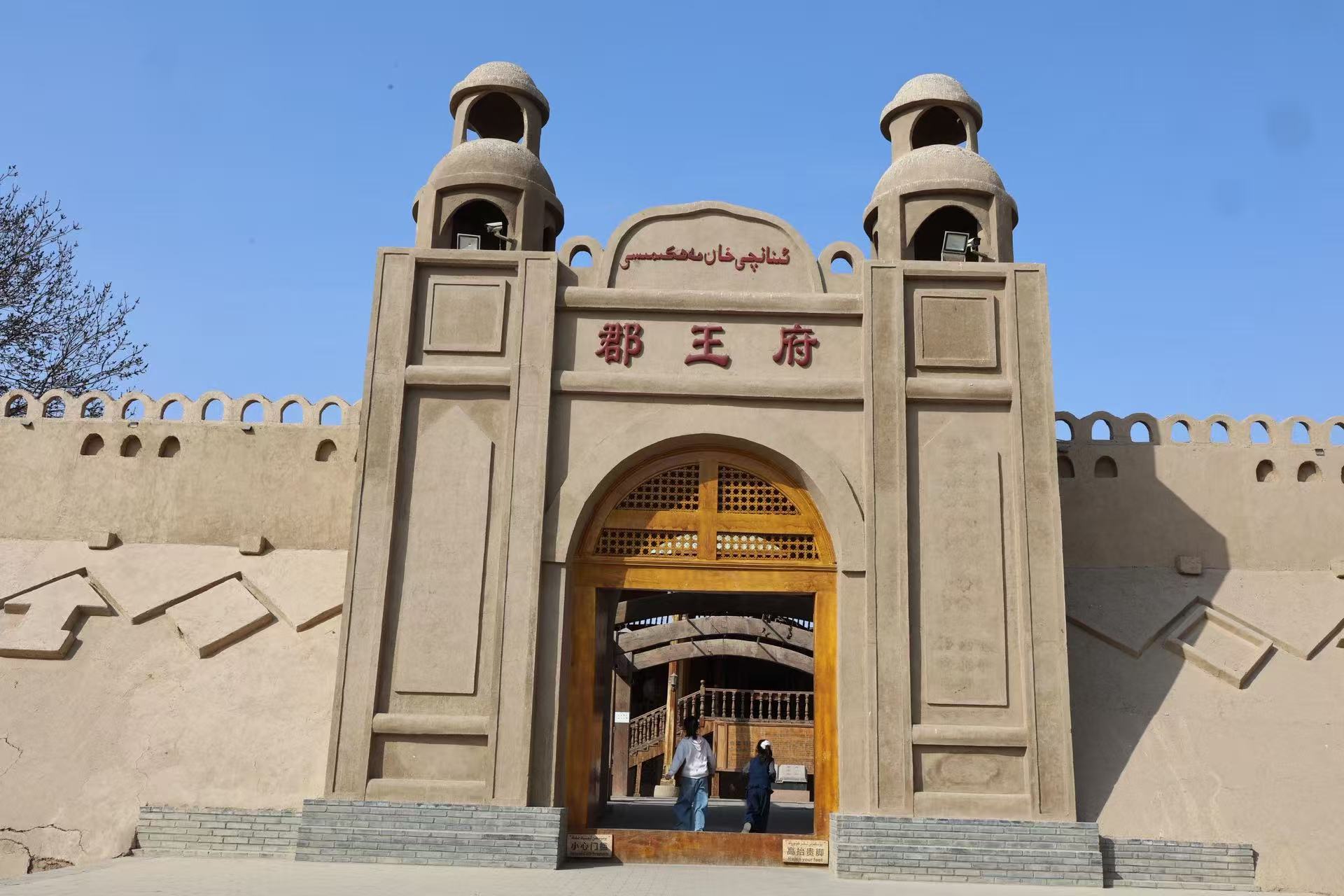 吐魯番郡王府正門為夯土風格建築,雙穹頂塔樓造型別致,門頭印有雙語 “郡王府” 標識,拱形木質大門典雅大氣,牆體搭配特色垛口,極具西域王府建築韻味 吐魯番郡王府正門為夯土風格建築,雙穹頂塔樓造型別致,門頭印有雙語 “郡王府” 標識,拱形木質大門典雅大氣,牆體搭配特色垛口,極具西域王府建築韻味